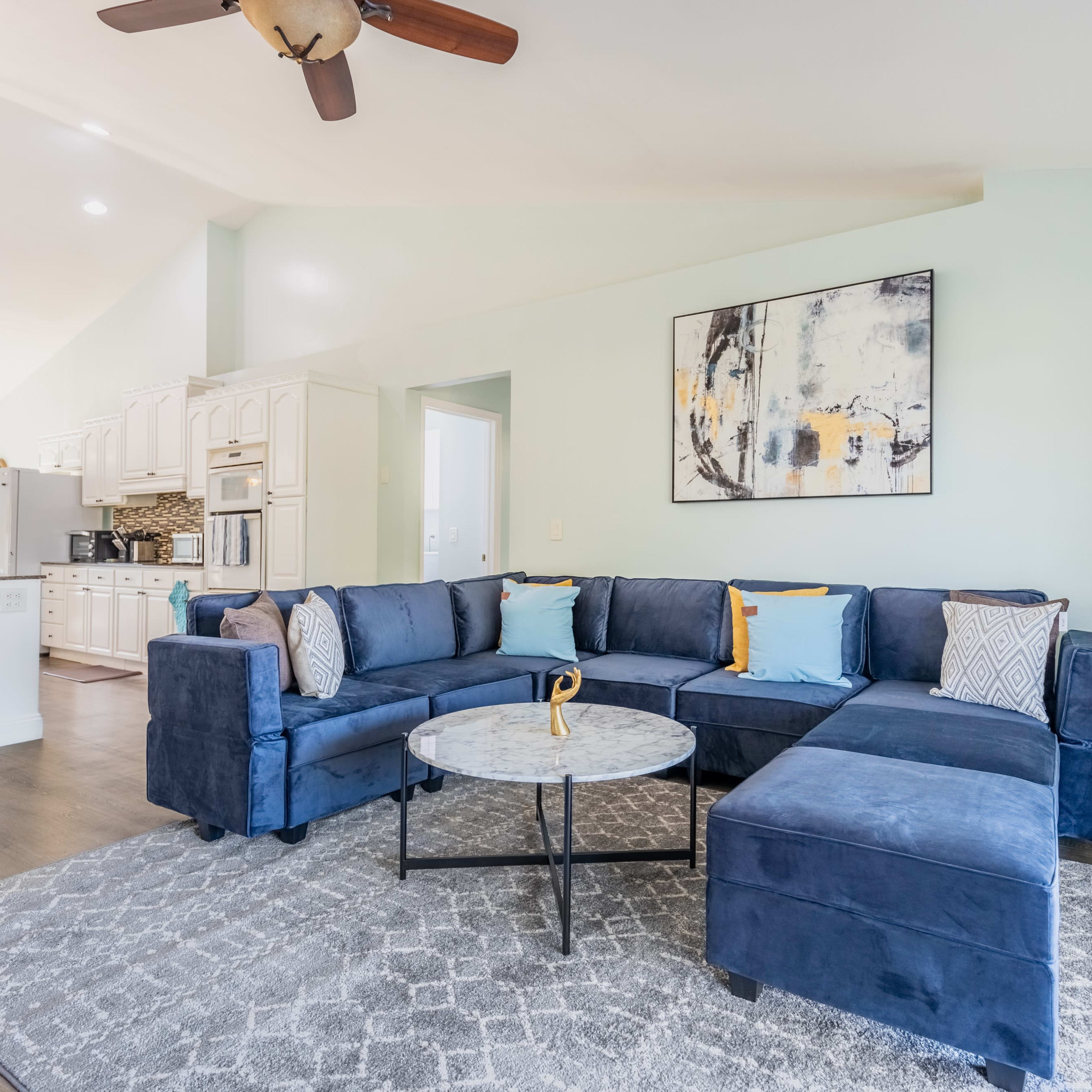 A blue sectional sofa with colorful cushions is arranged around a circular coffee table on a patterned rug in a light-filled living area that opens to a kitchen.