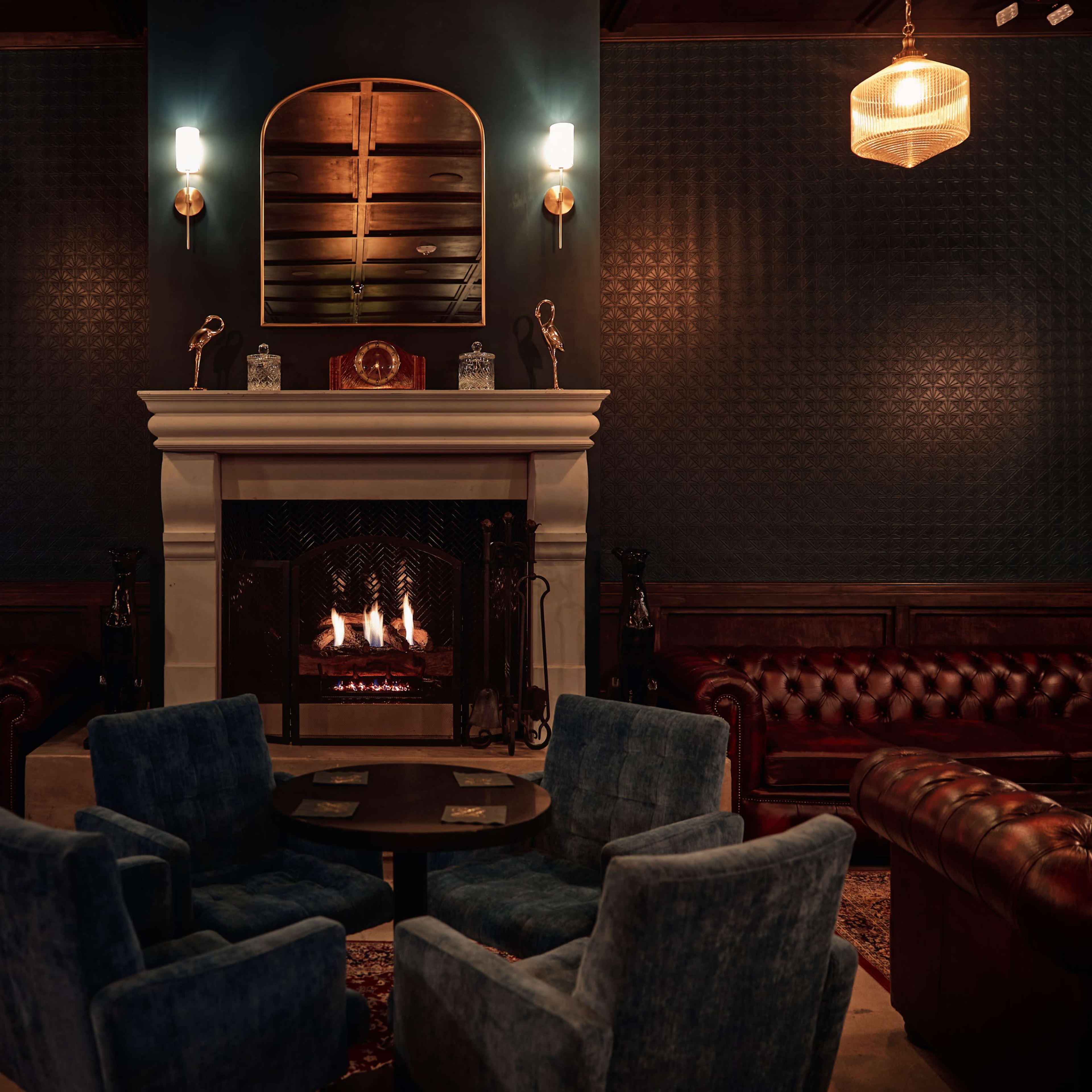 A cozy lounge features a fireplace surrounded by leather sofas and blue velvet chairs under warm lighting.