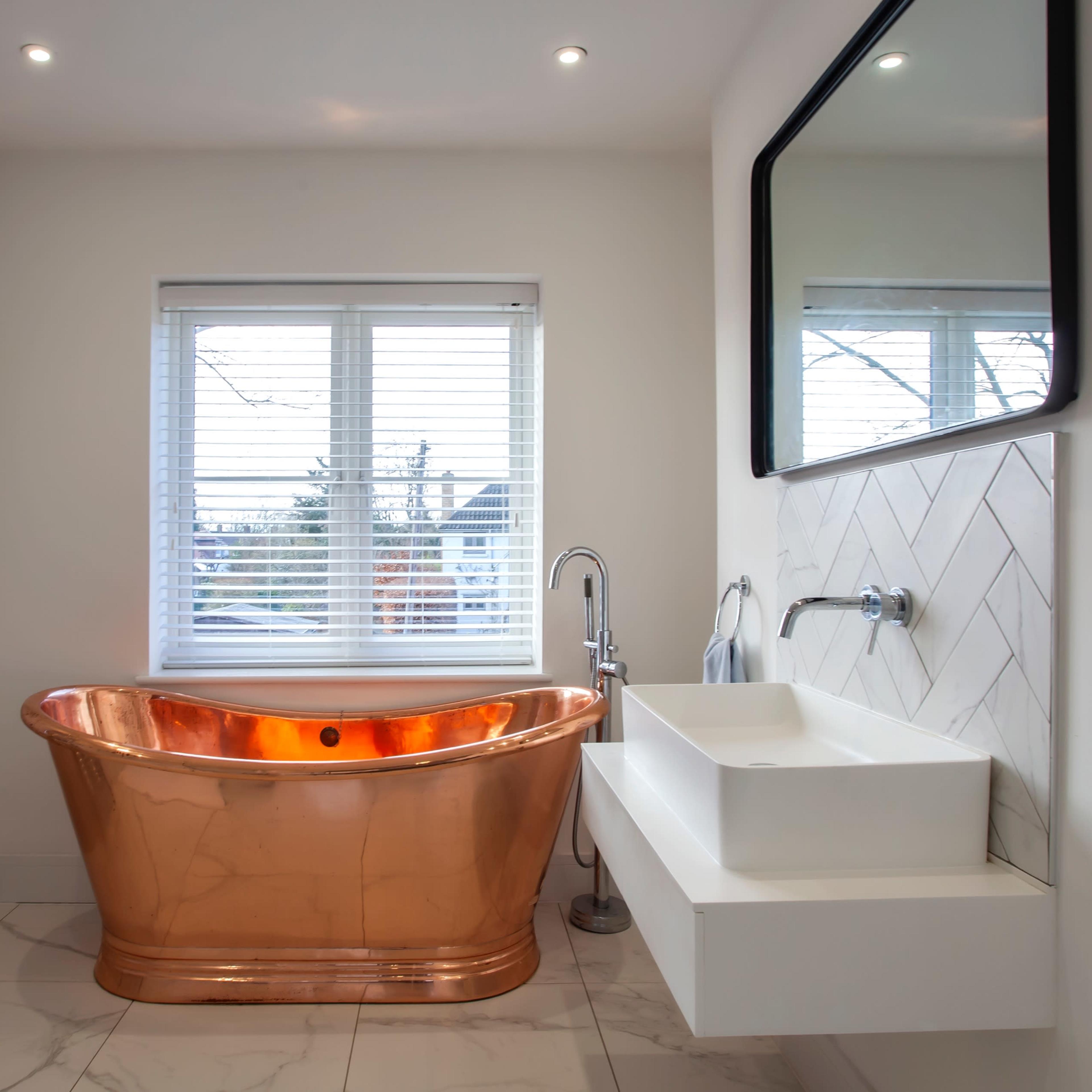 A modern bathroom featuring a copper freestanding bathtub, a wall-mounted sink, and a large mirror, with light coming in through a window.