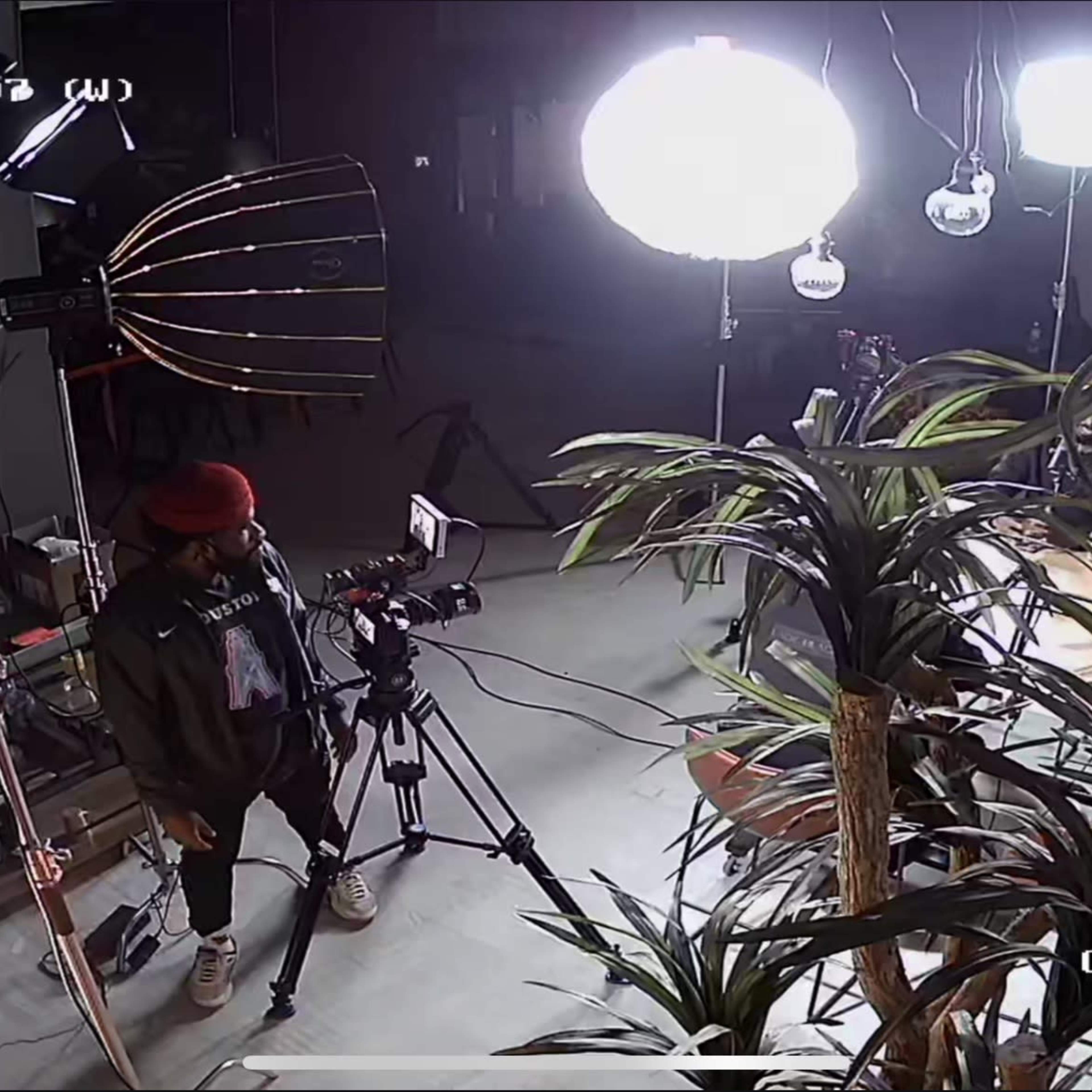 A person standing with a camera near studio lighting and various decorative plants in an indoor setting.