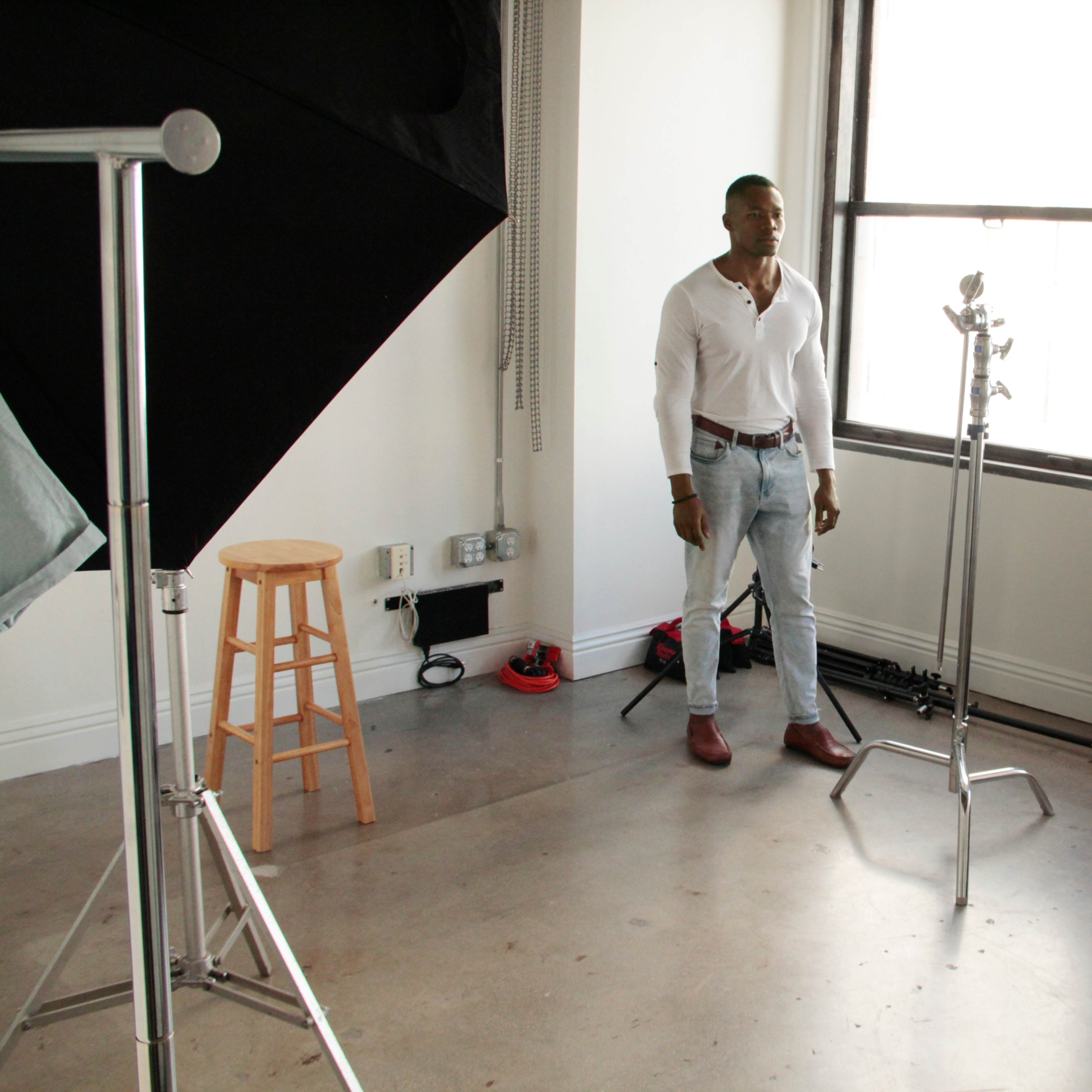 A man stands in a photography studio filled with equipment, including a stool, light stands, and a reflector.