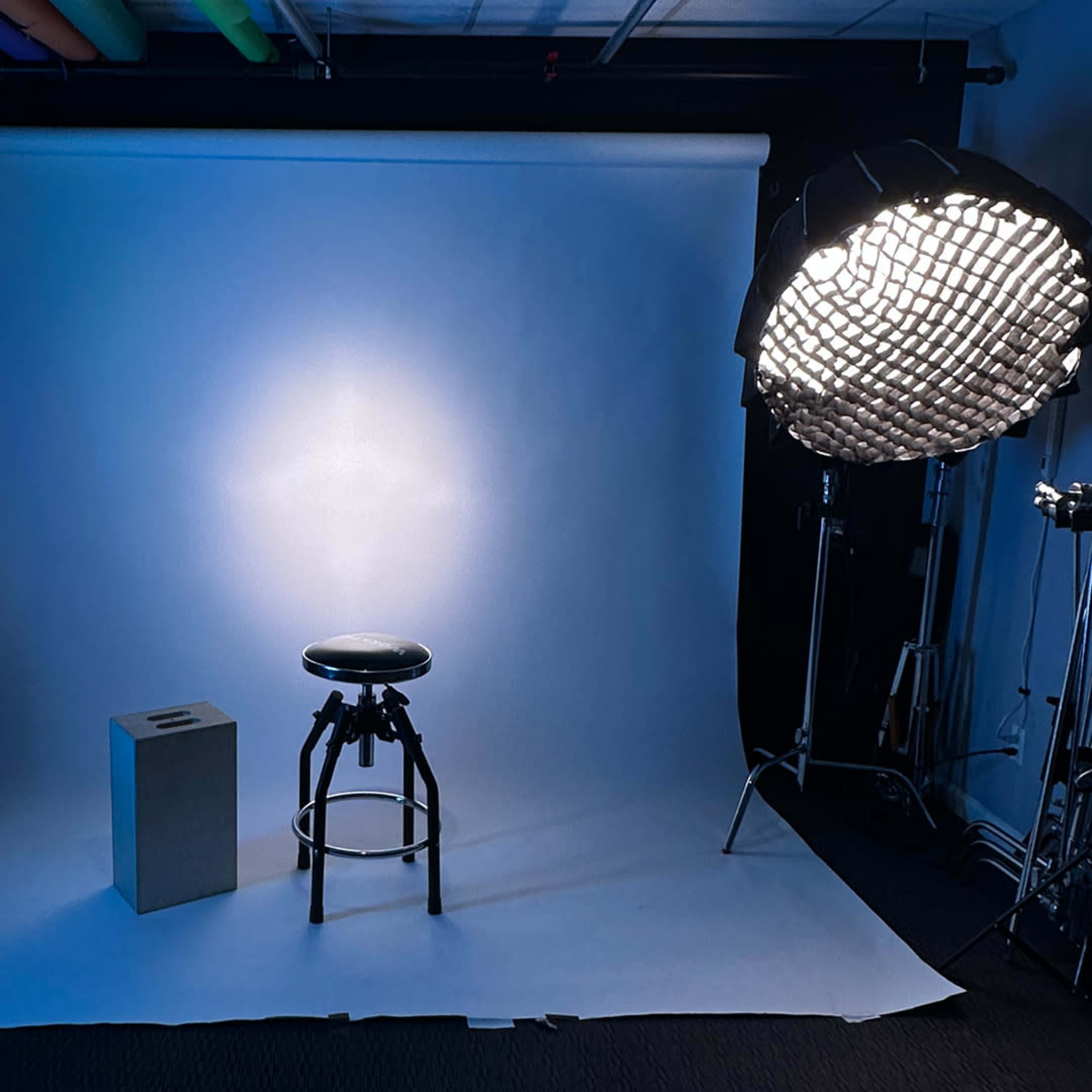 A photography studio setup featuring a black stool and a gray cube in front of a white backdrop, illuminated by a large softbox and accented by colorful LED lights on the side.