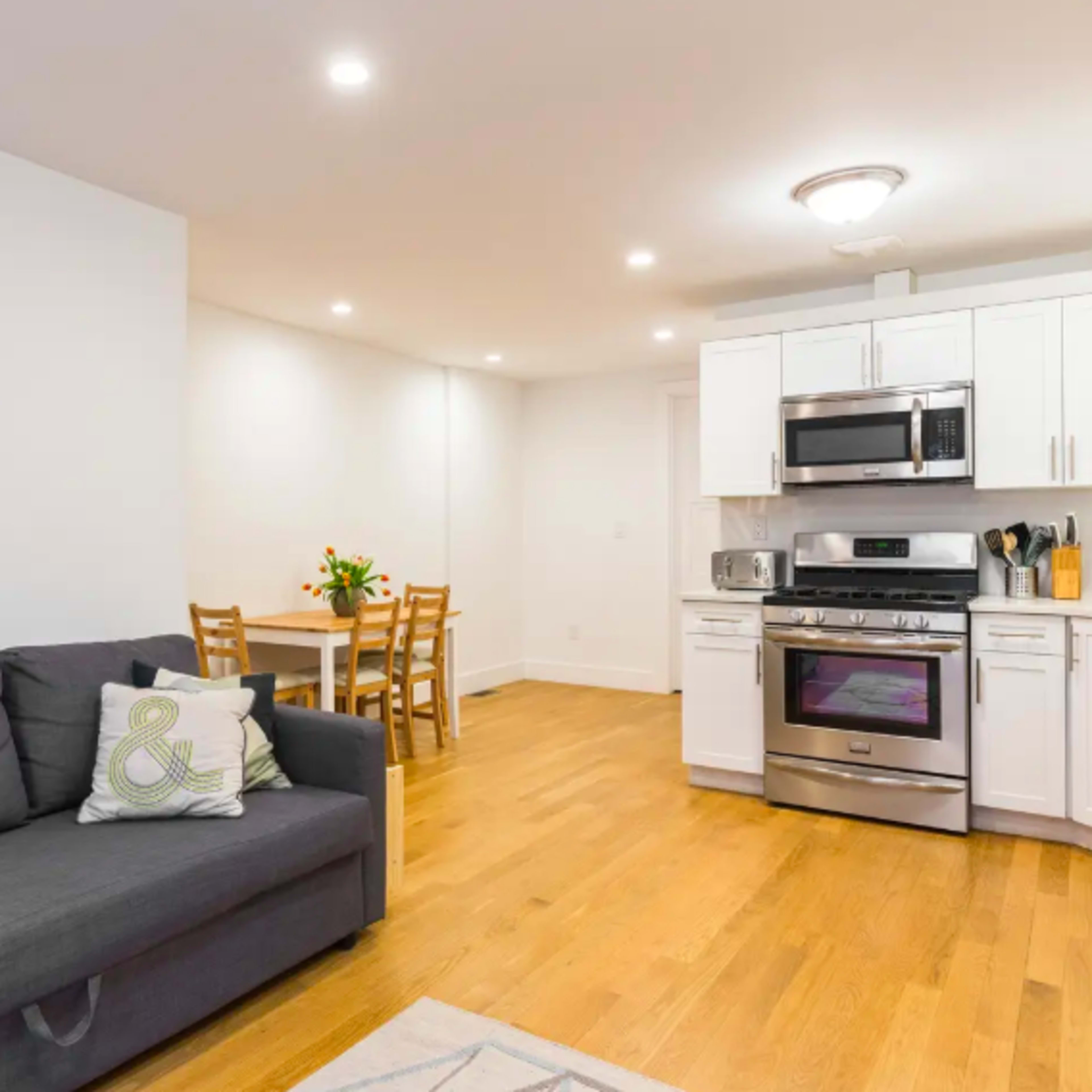 A modern kitchen and living area with a gray sofa, a dining table with chairs, and stainless steel kitchen appliances.