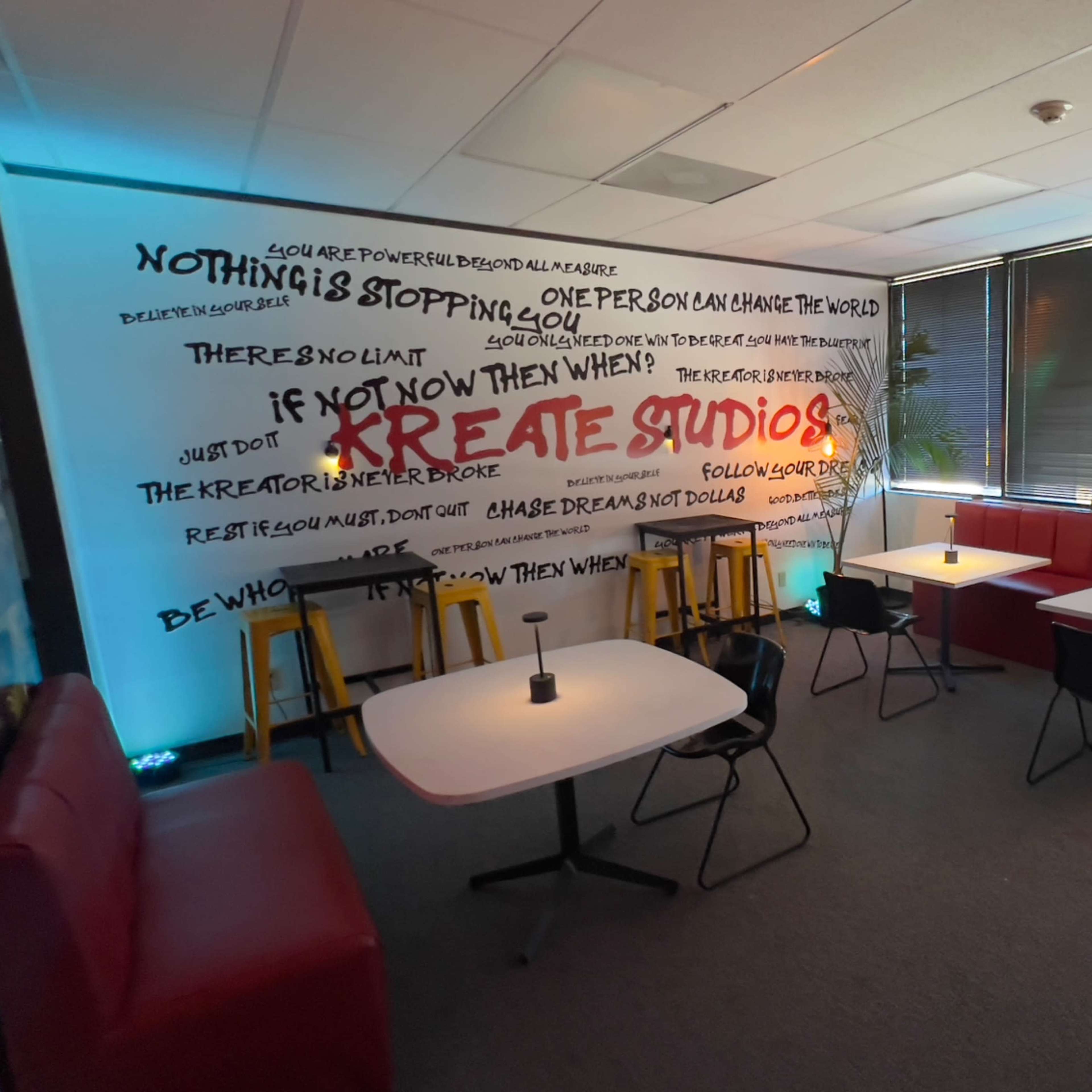 A creative workspace features white tables, chairs, and a large wall mural with motivational text, surrounded by bright lighting and greenery.