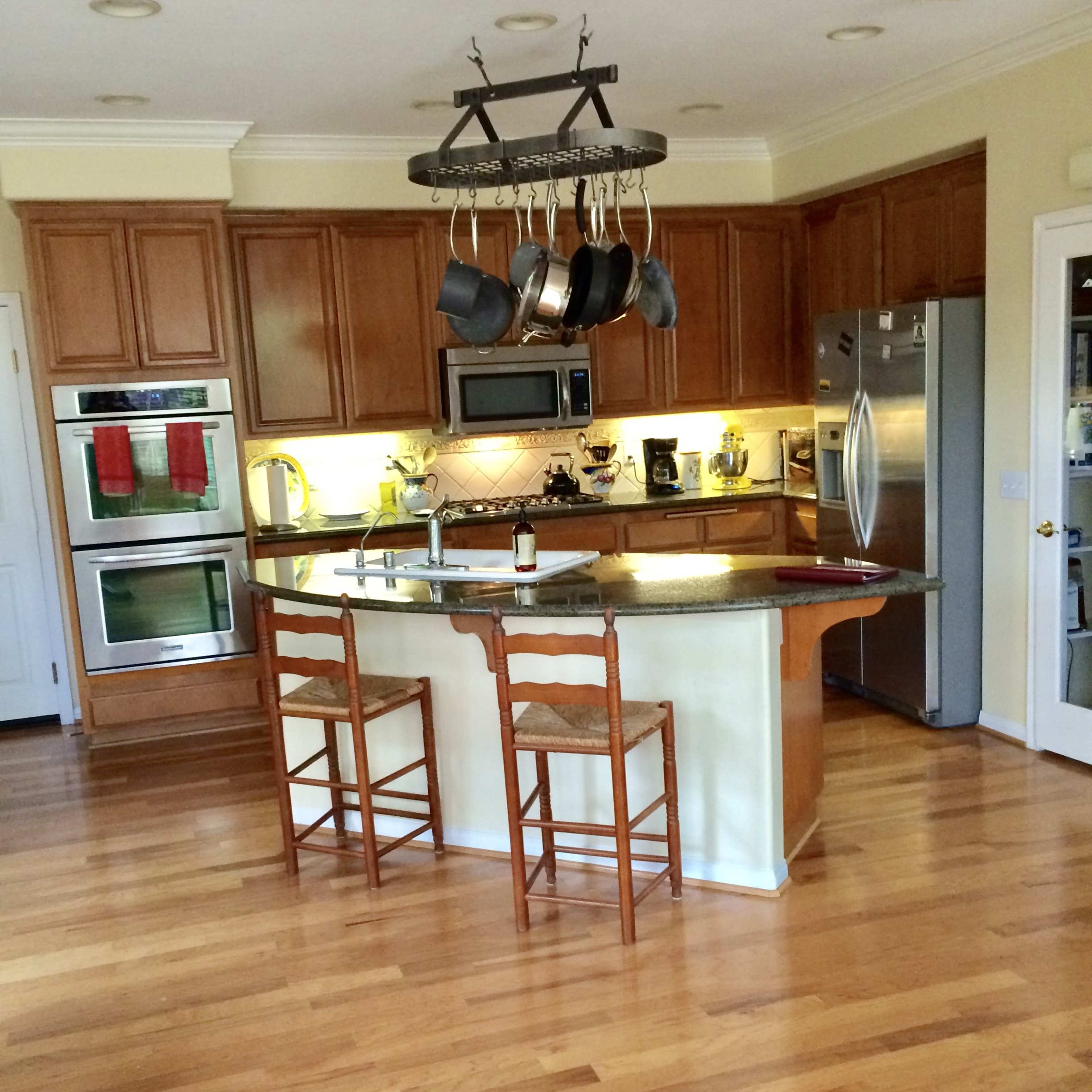A modern kitchen features wooden cabinets, stainless steel appliances, a central island with two wooden stools, and a pot rack hanging overhead.