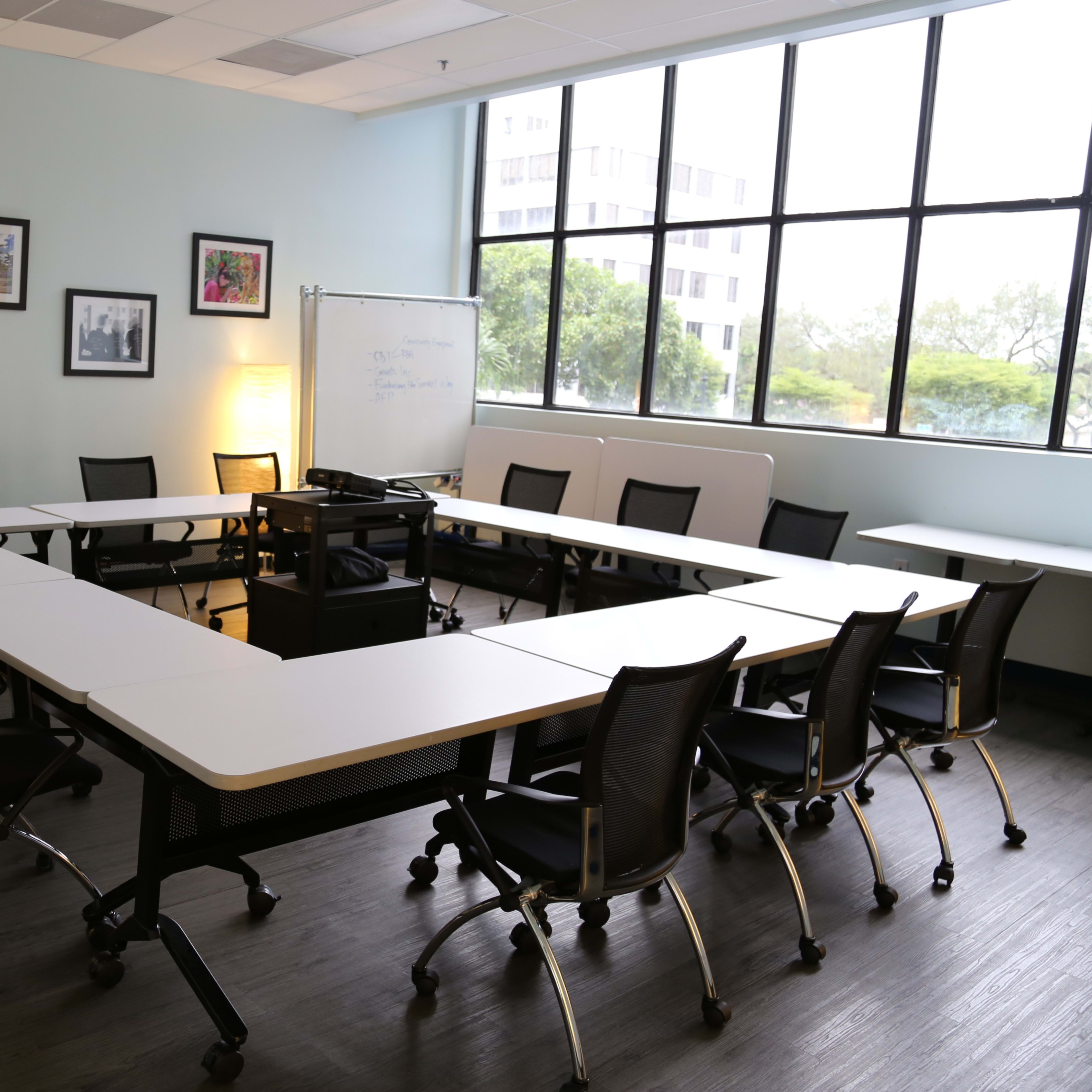 A well-lit conference room features a rectangular arrangement of tables and chairs, with large windows providing natural light.