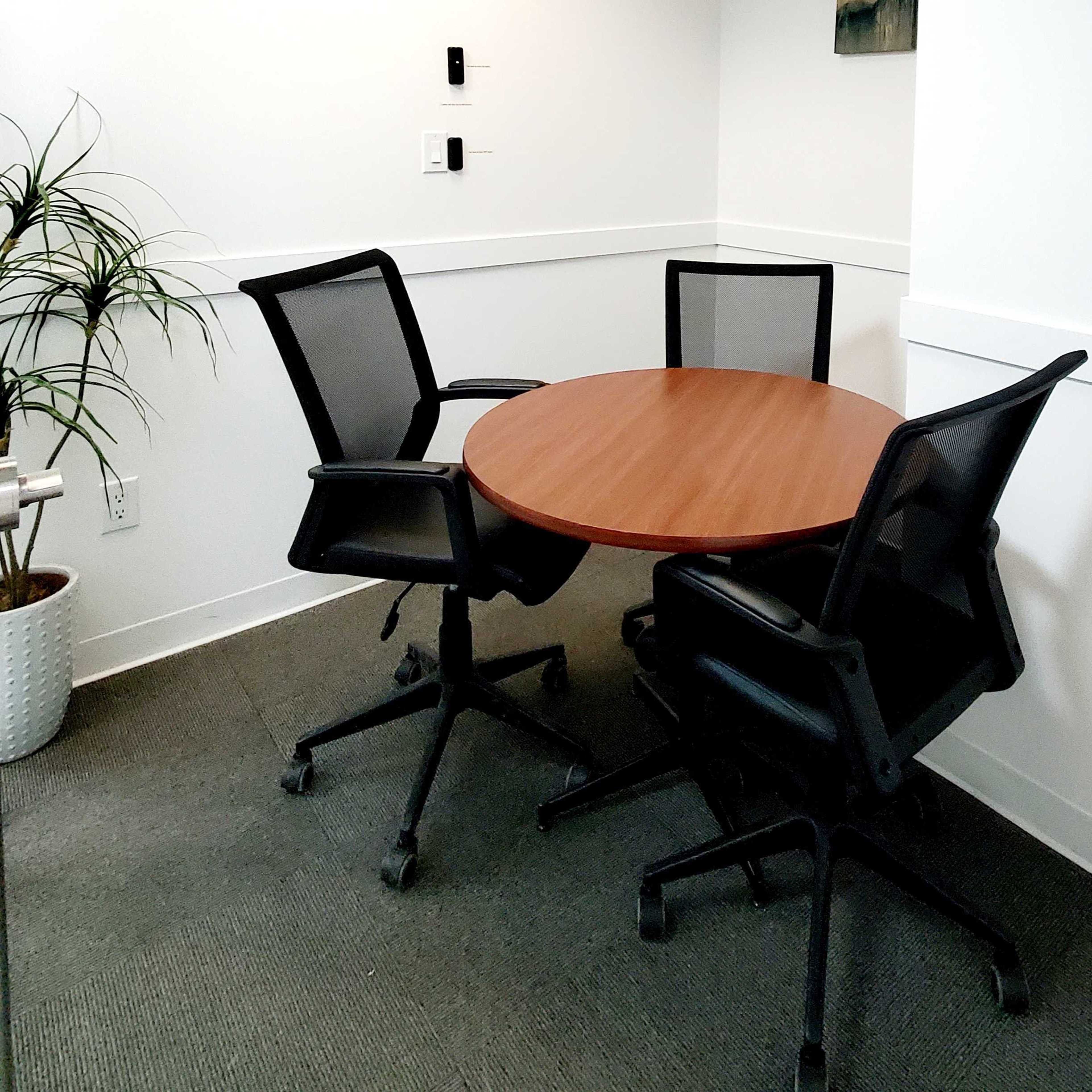 A round wooden table is positioned in a corner of a room with three black mesh chairs around it.