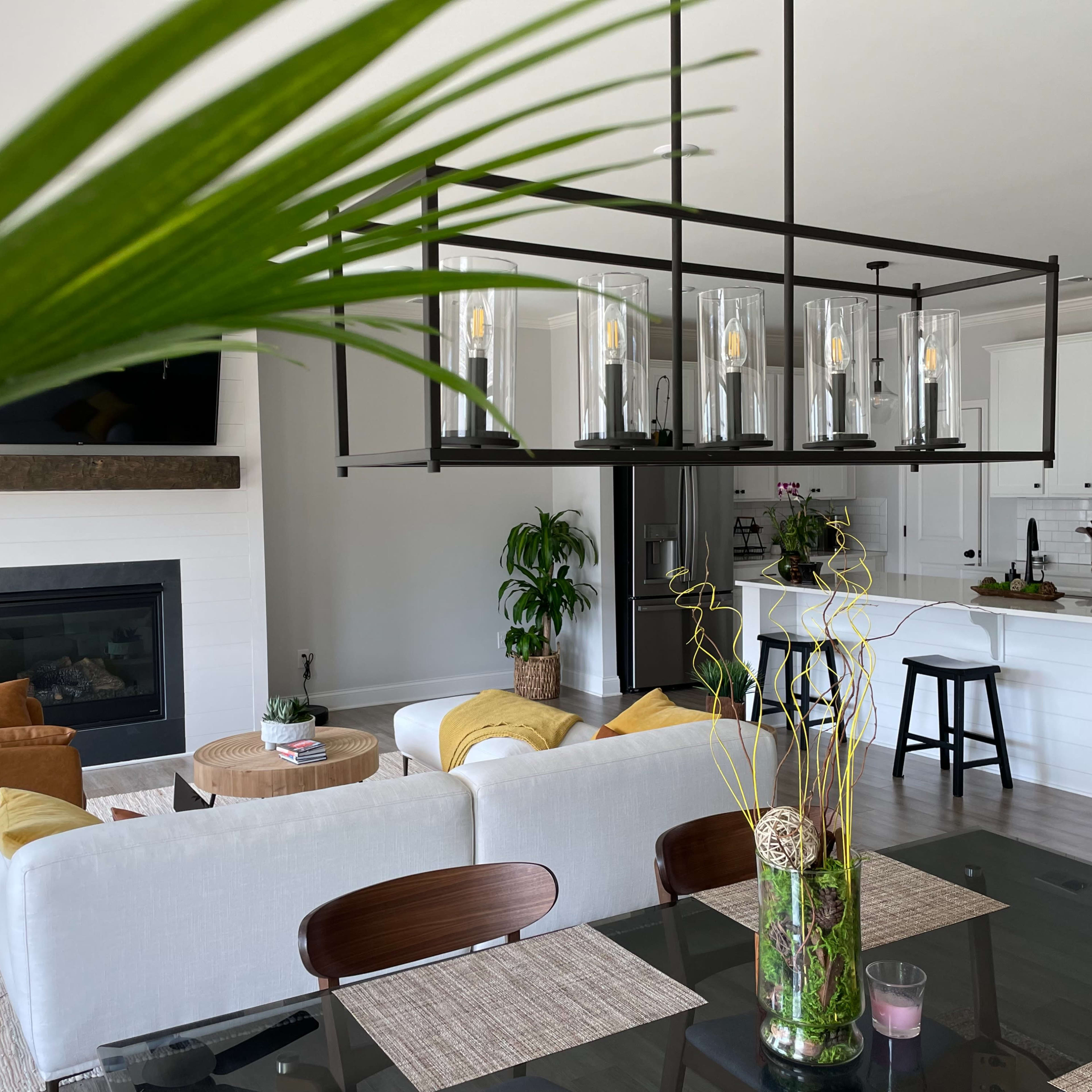 A modern living and dining area features a large chandelier above a dark table, with a beige sofa, brown leather chairs, and a fireplace in the background.