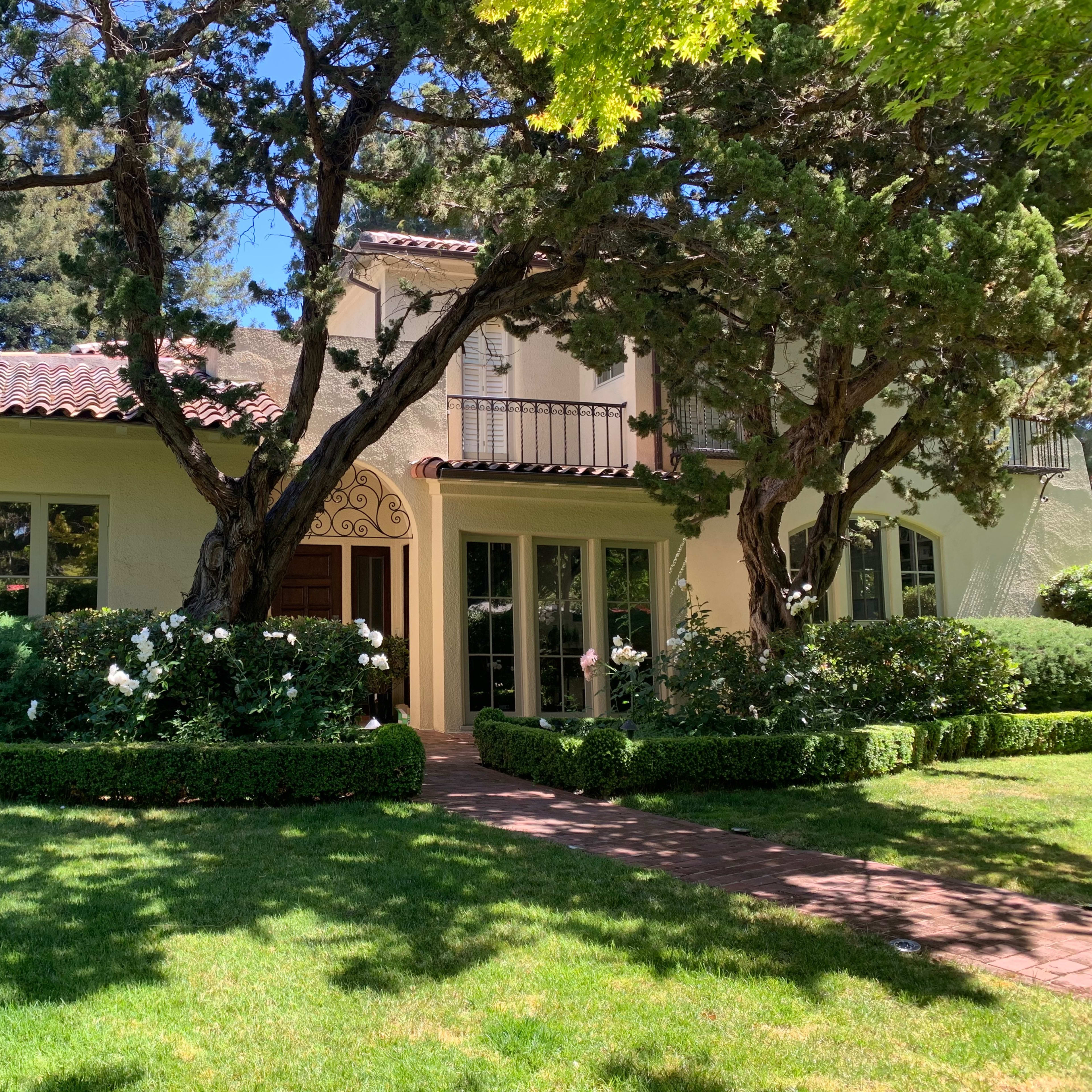 A two-story house with a tiled roof, surrounded by neatly trimmed hedges and a lawn, under a clear blue sky.
