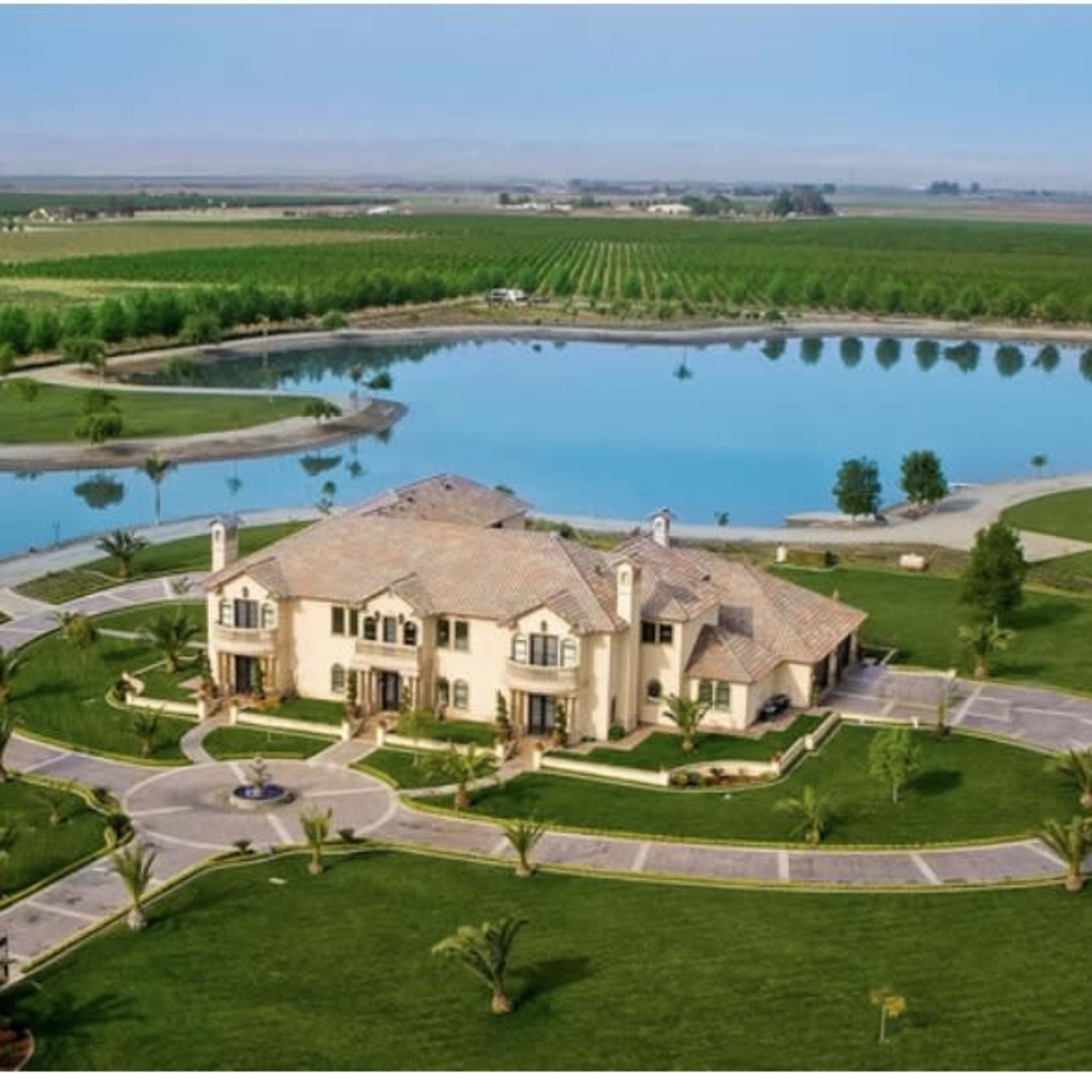 A large estate with a circular driveway, surrounded by well-manicured lawns, palm trees, and a reflective lake, set against a backdrop of agricultural fields.