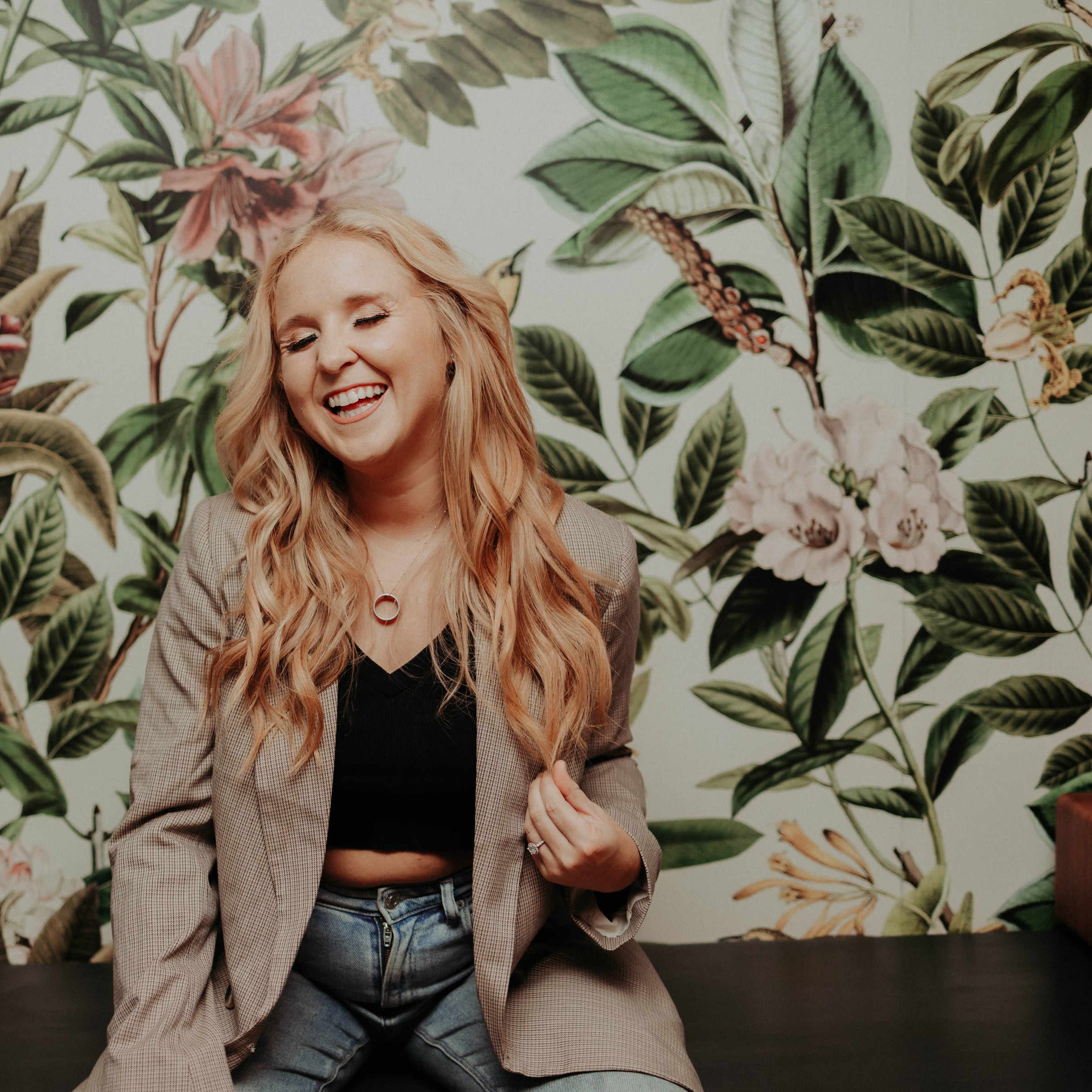 A woman with long blonde hair sits in front of a floral wallpaper, wearing a blazer and jeans, smiling as she poses.