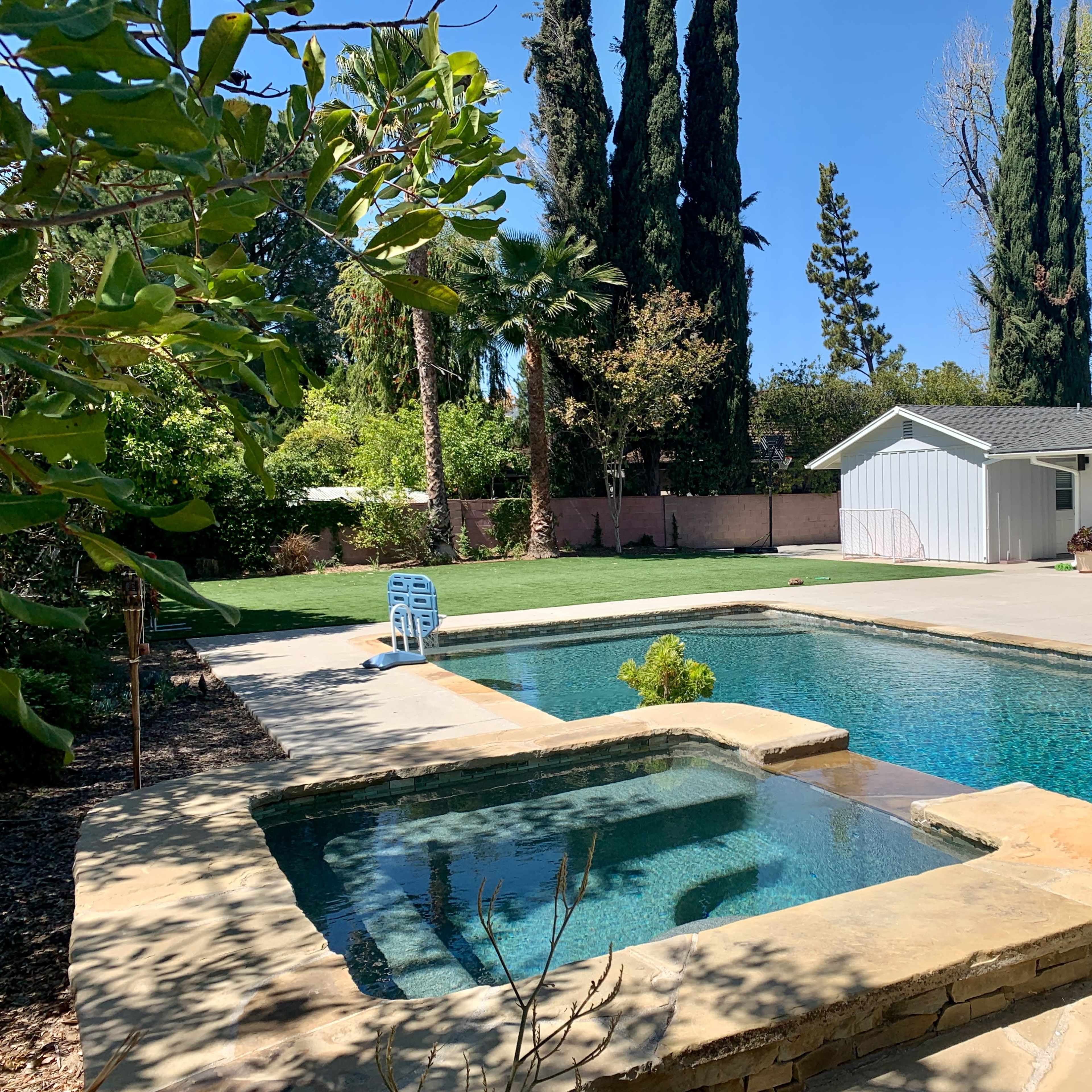 A swimming pool with a spa, surrounded by greenery and a single-story house in the background.