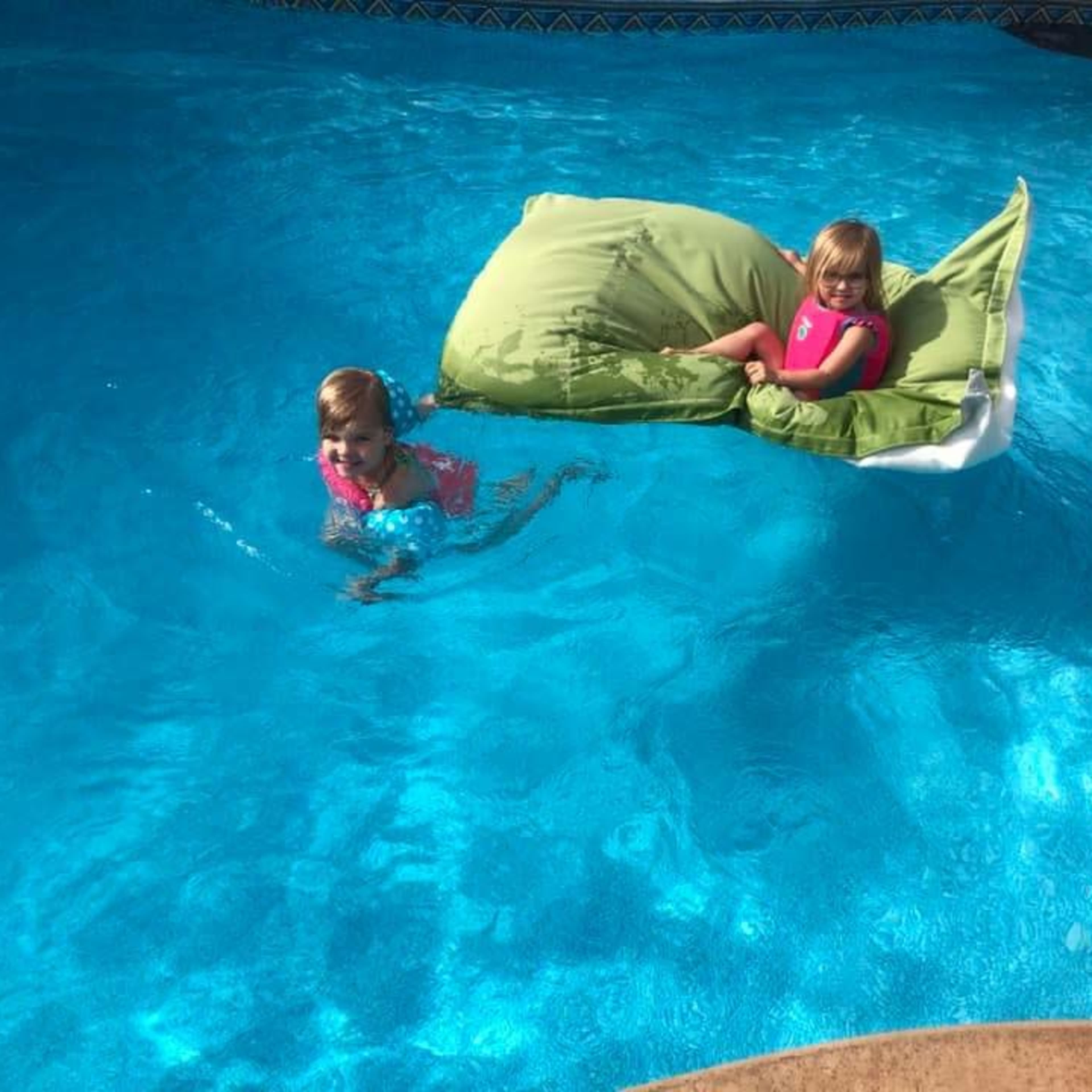 Two girls are swimming in a pool, with one on a green float and the other in the water nearby.