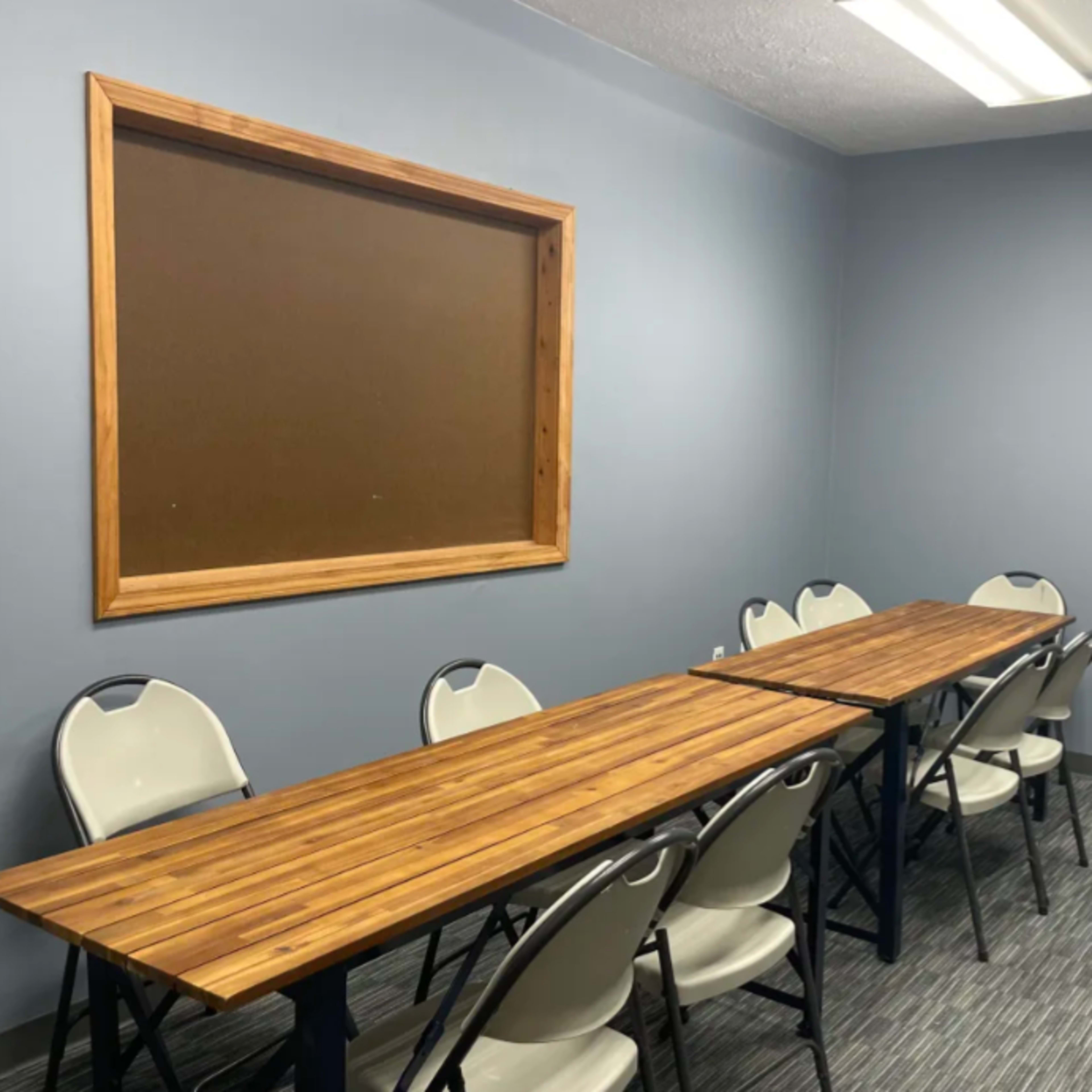 A small meeting room featuring a long wooden table and several plastic chairs against a blue wall with a bulletin board.