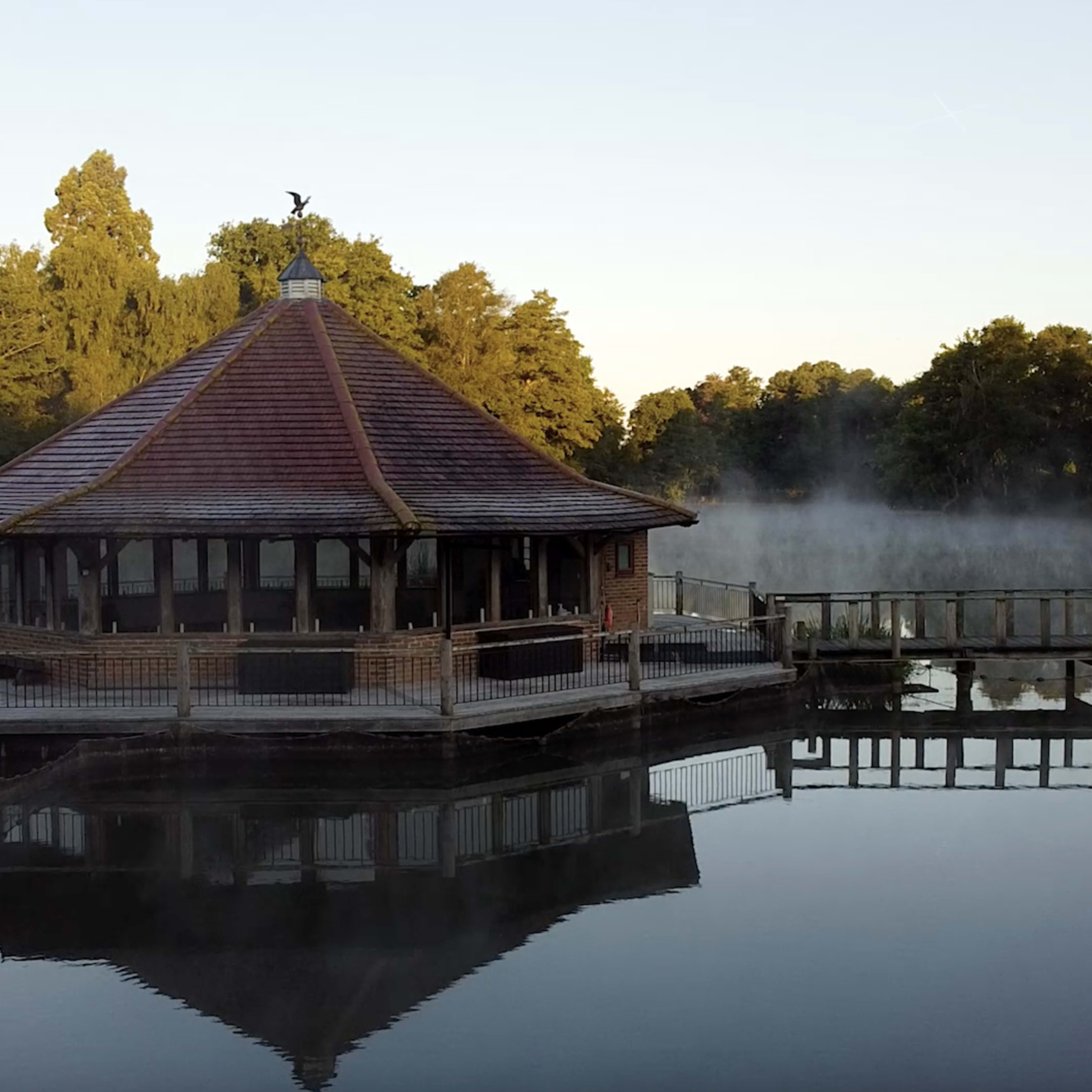 A wooden gazebo with a conical roof sits on the edge of a calm, misty lake, surrounded by trees and reflected in the water.