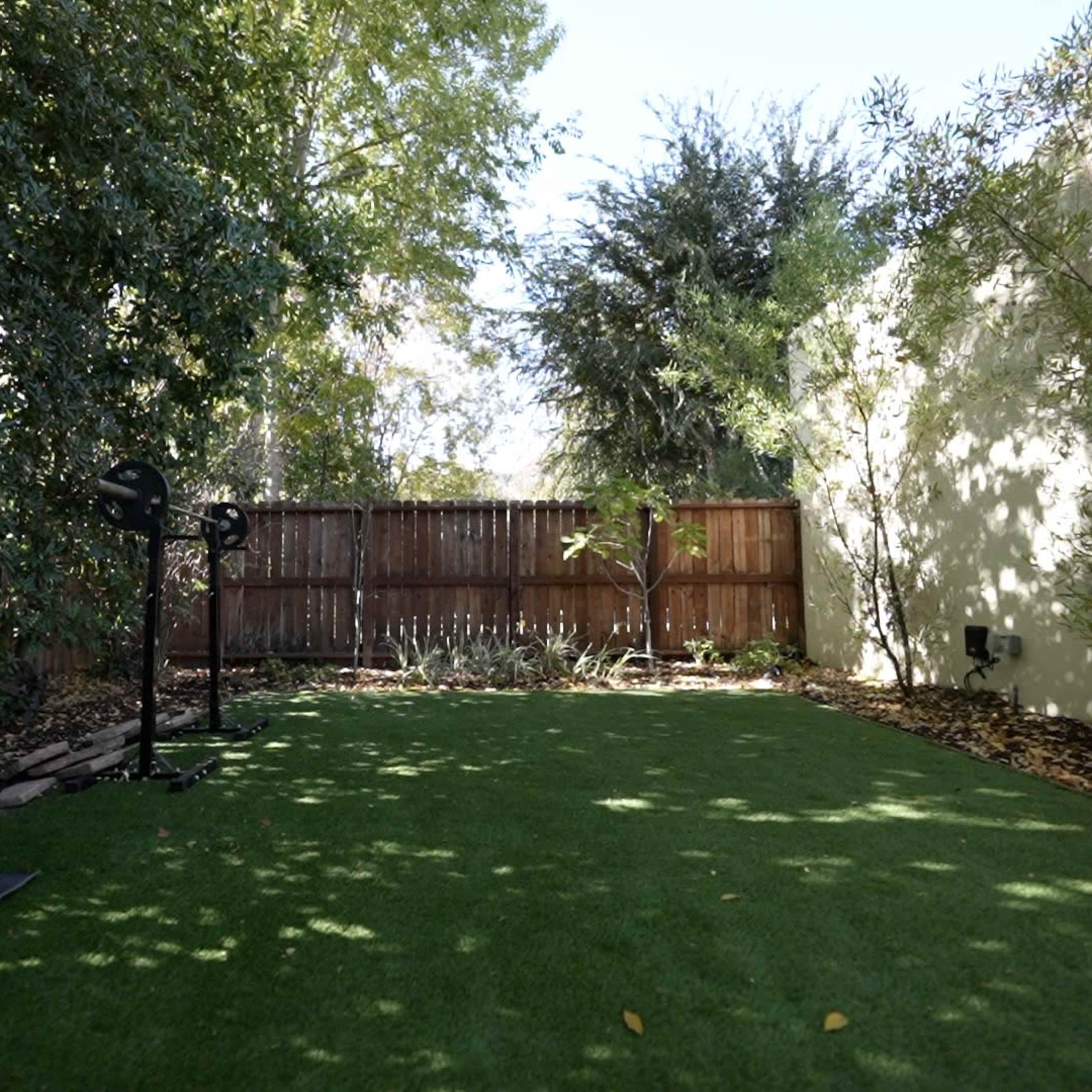 A fenced backyard with artificial grass, surrounded by trees and shrubs, featuring a basketball hoop in one corner.