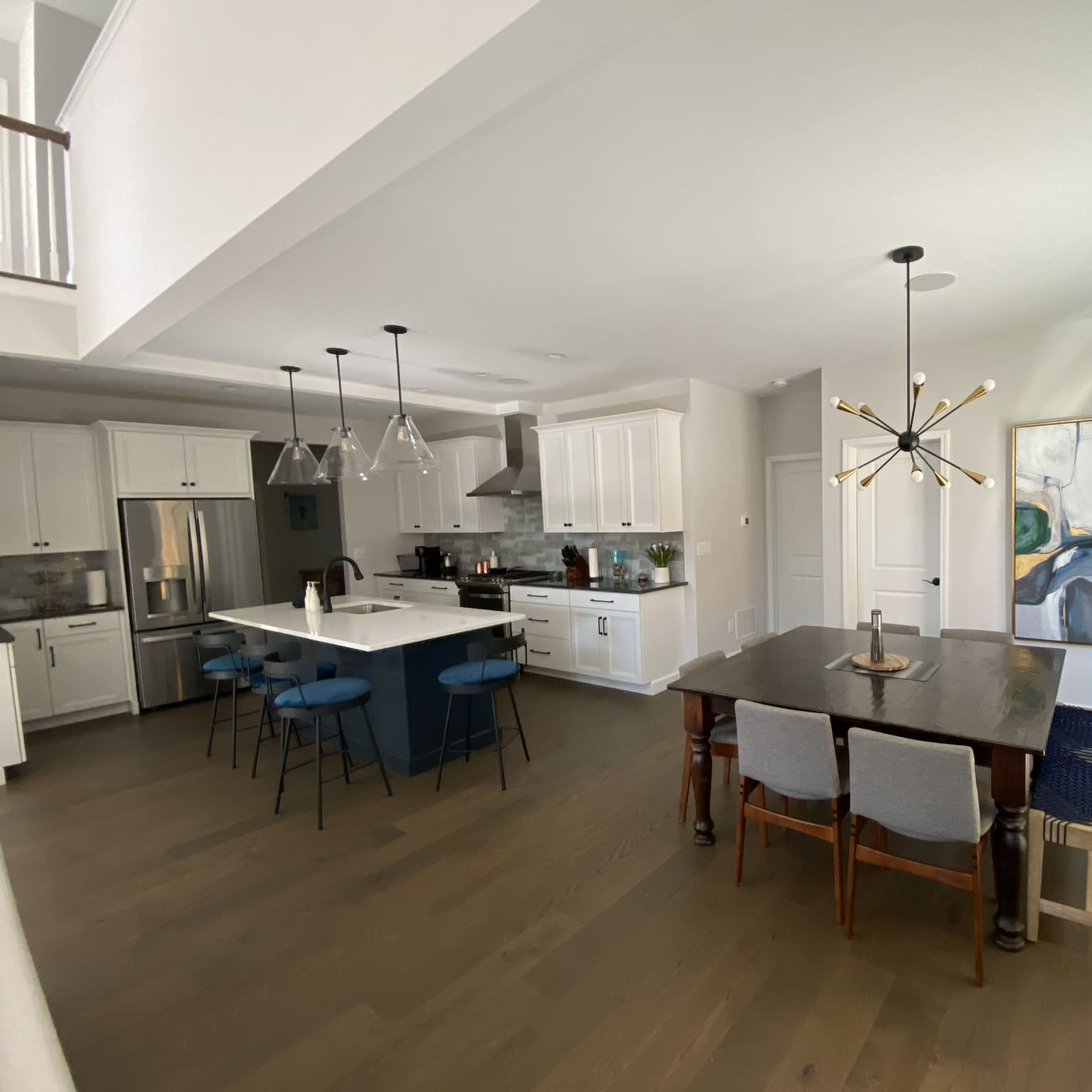 A modern kitchen and dining area with a centrally placed table, blue bar stools, and large windows allowing natural light to enter.