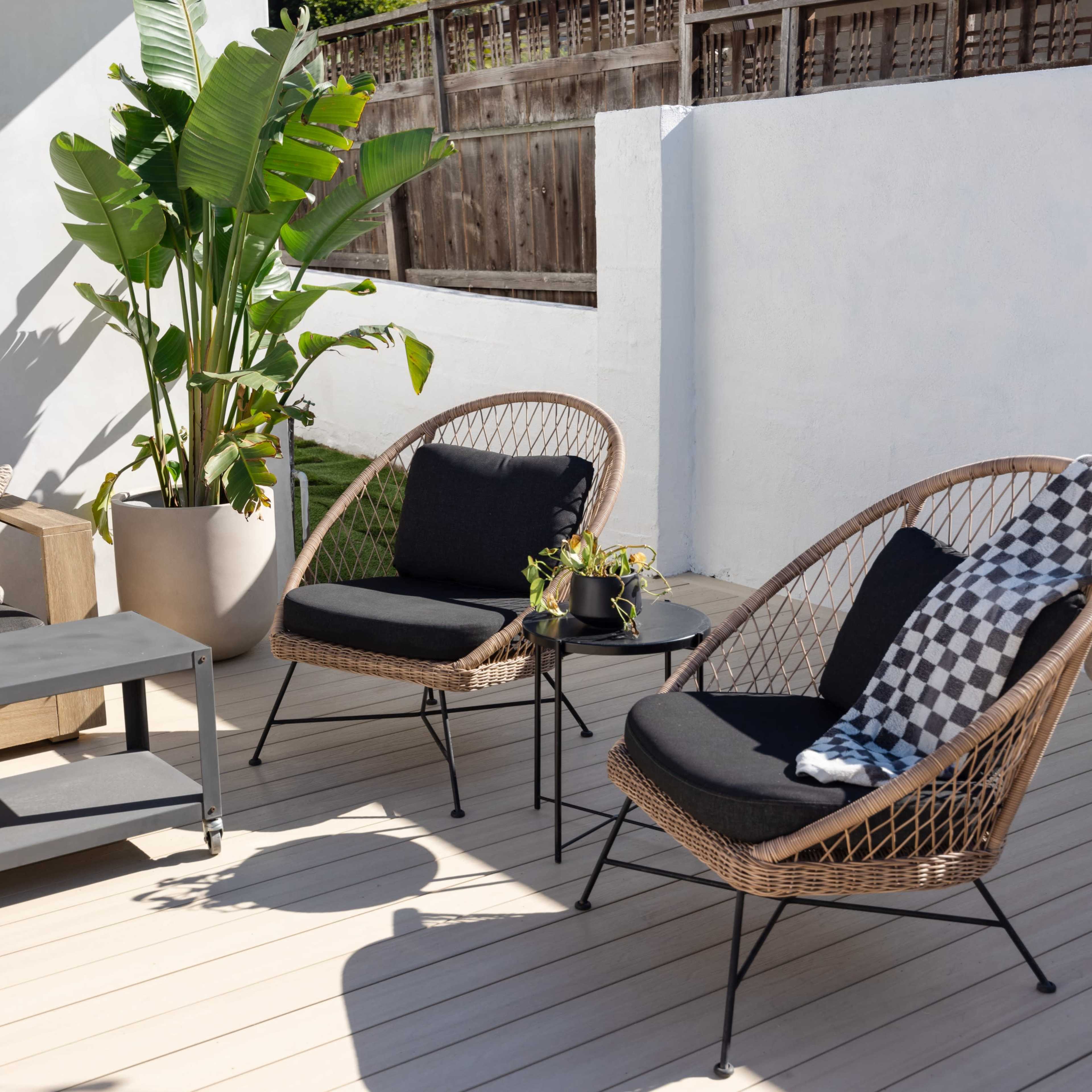 A modern outdoor patio featuring two wicker chairs, a small table, a large potted plant, and a sofa against a light-colored wall.