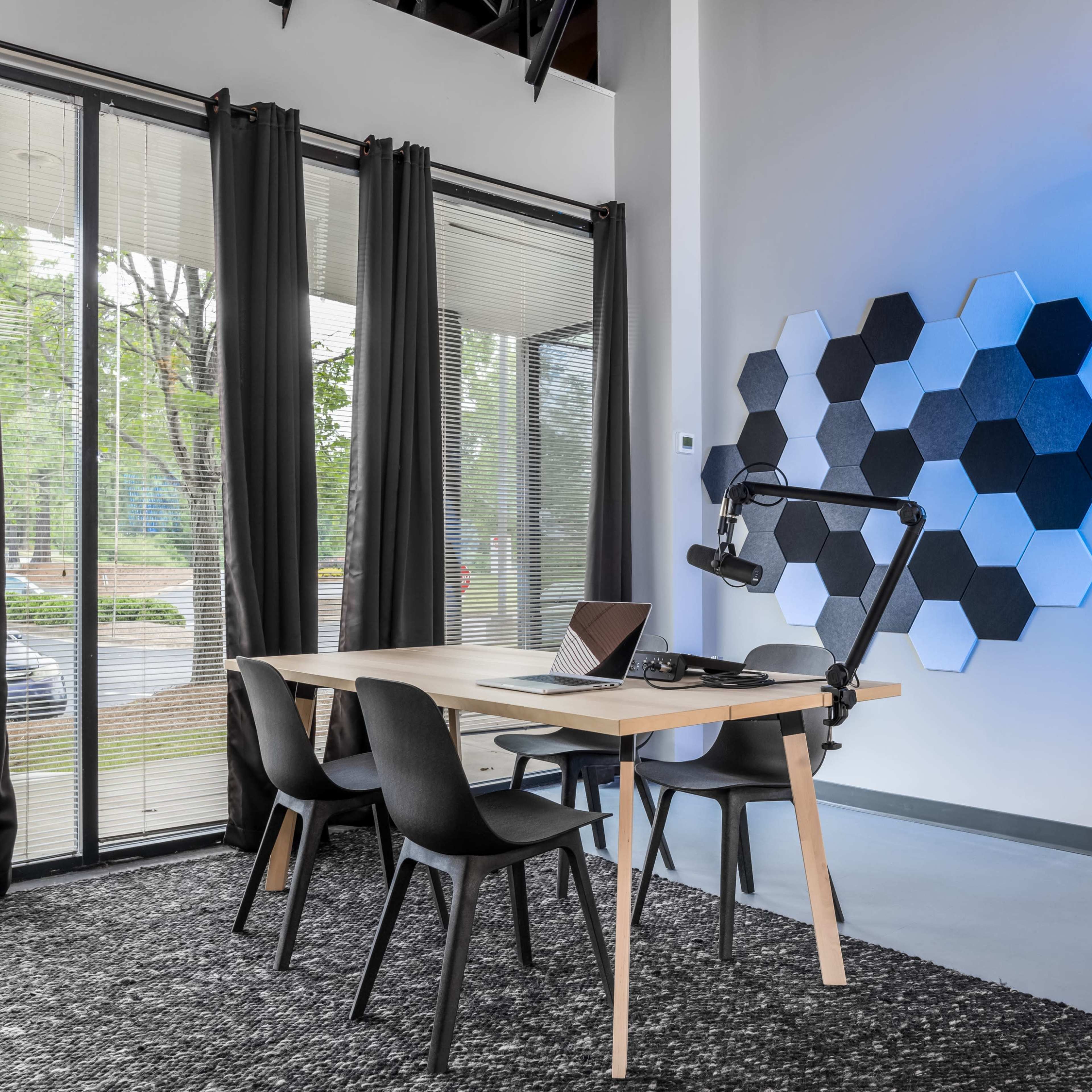 A modern workspace features a wooden table with a laptop and an adjustable lamp, surrounded by four black chairs, against a wall with hexagonal decorations and large windows draped with curtains.