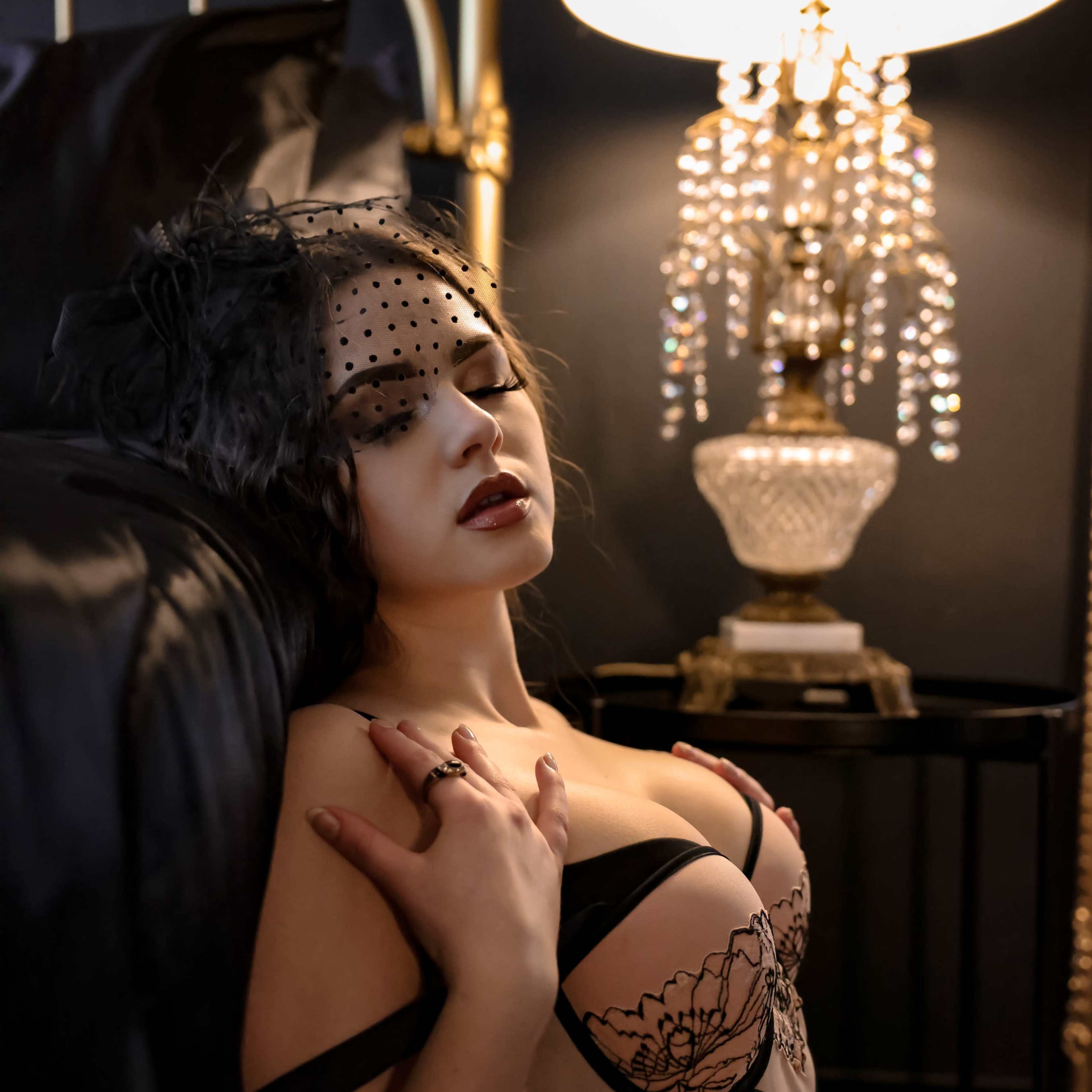 A woman in lingerie poses near a lamp adorned with crystals, resting against a black headboard.