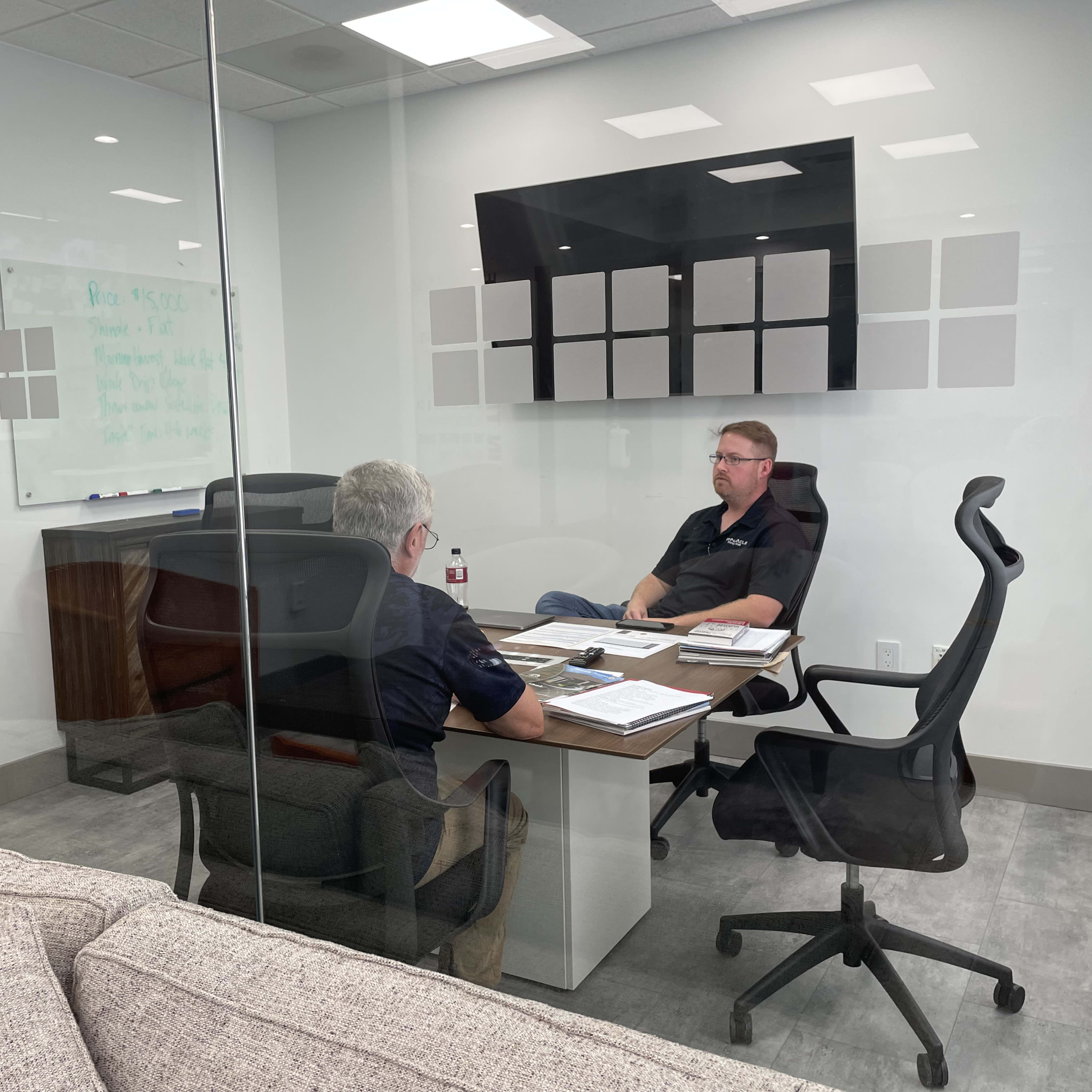 Two men are seated at a conference table in a glass-walled meeting room, discussing documents and notes.