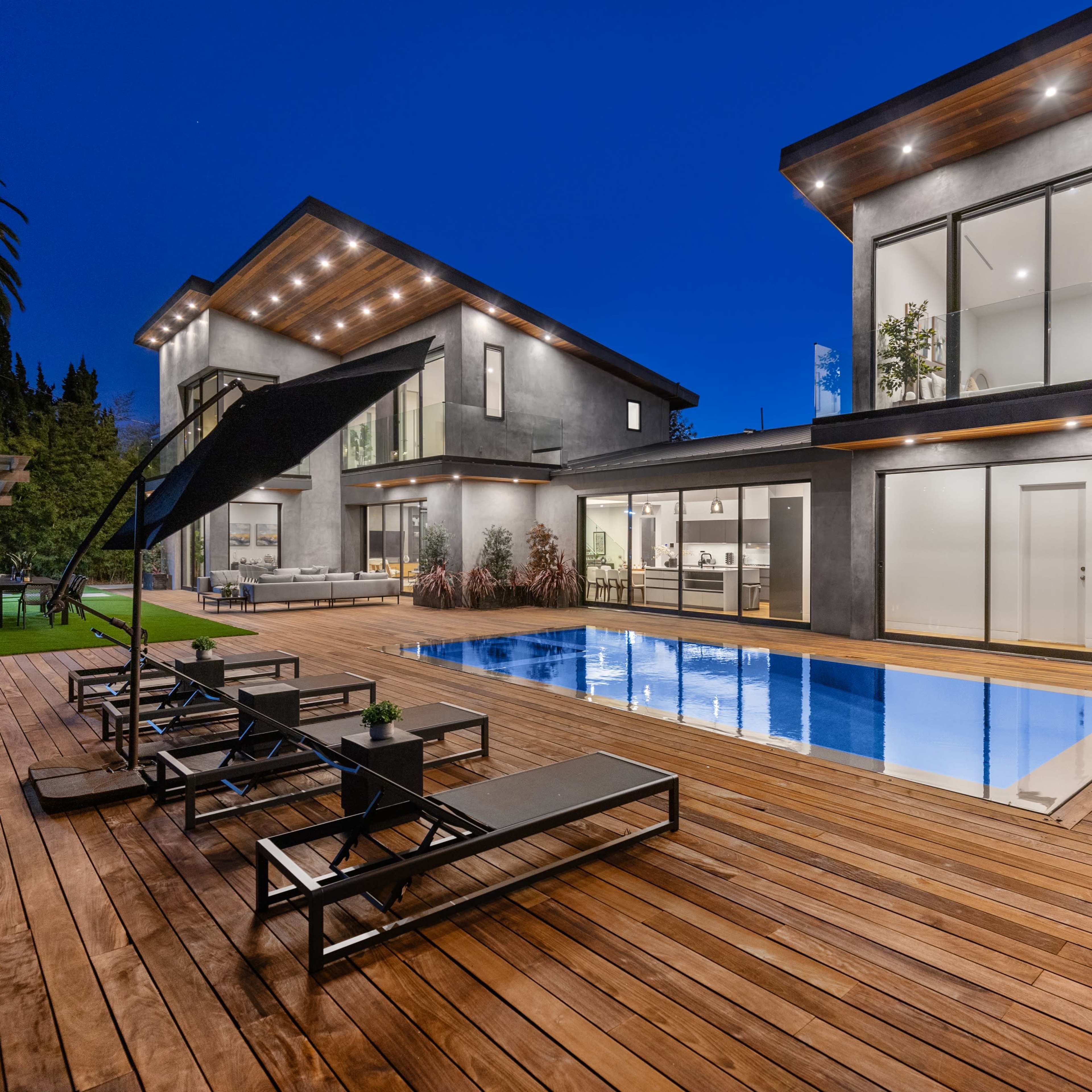 A modern house with large windows, a swimming pool surrounded by wooden decking, and lounge chairs under a large umbrella, set against a twilight sky.