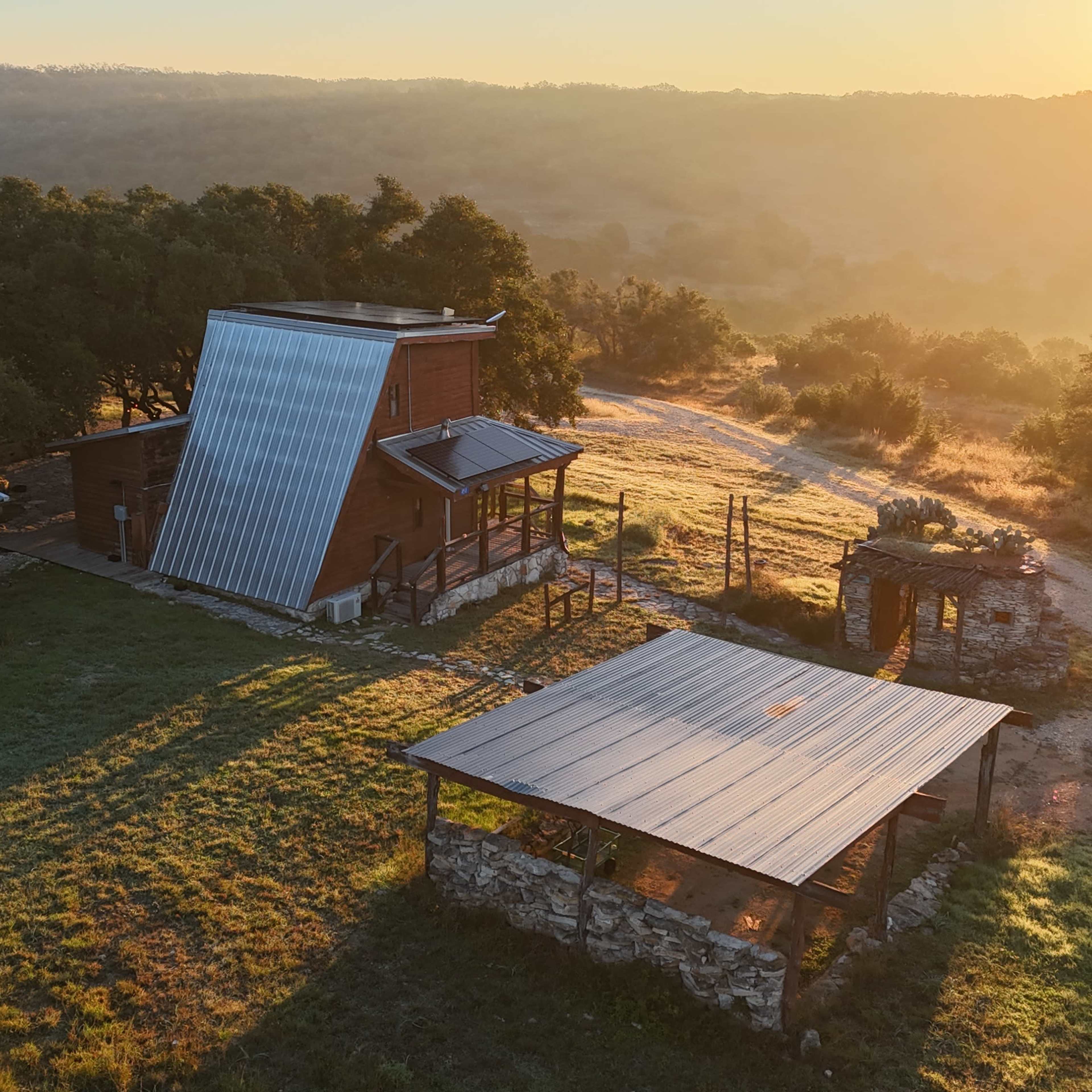 A rustic cabin with a metal roof and solar panels, surrounded by a grassy landscape bathed in warm sunlight during sunrise.