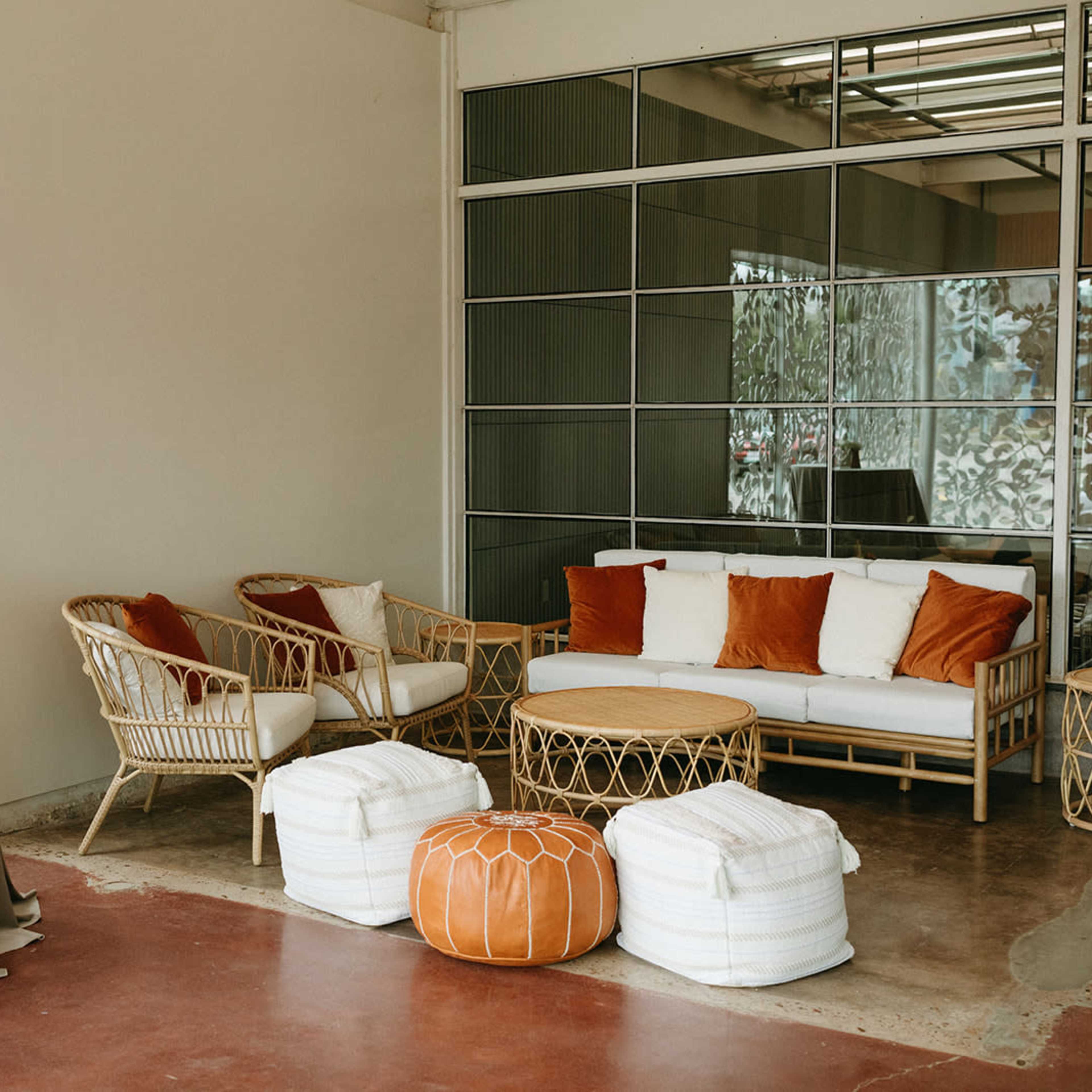 A cozy seating area features a sofa with orange and white pillows, two rattan chairs, a wooden coffee table, and poufs, set against a glass wall.