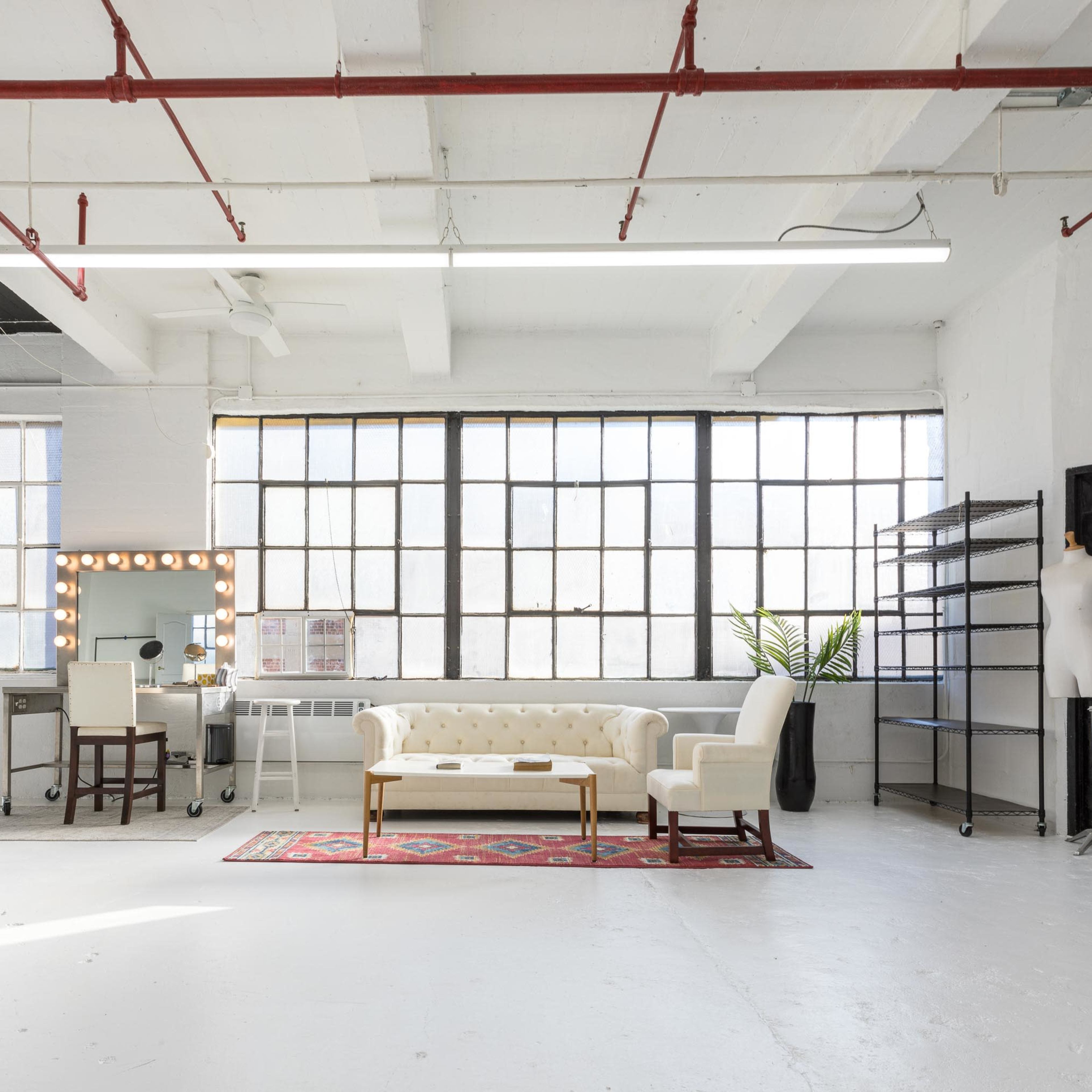 A spacious room features large windows, a tufted white sofa, a decorative mirror with lights, a metal shelving unit, and a plant.