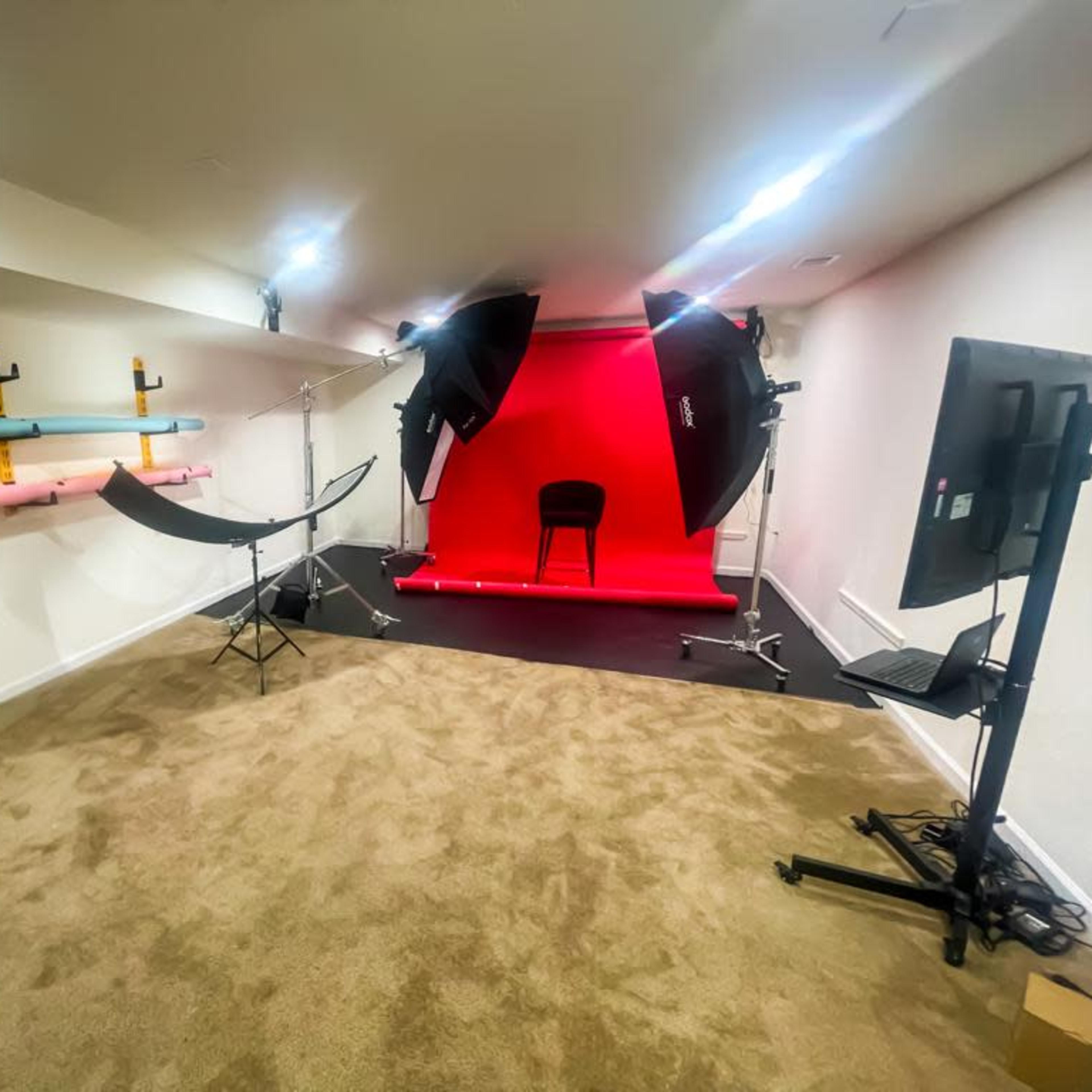 A photography studio featuring a red backdrop, studio lights, a chair, and various colored backdrop rolls on the wall.