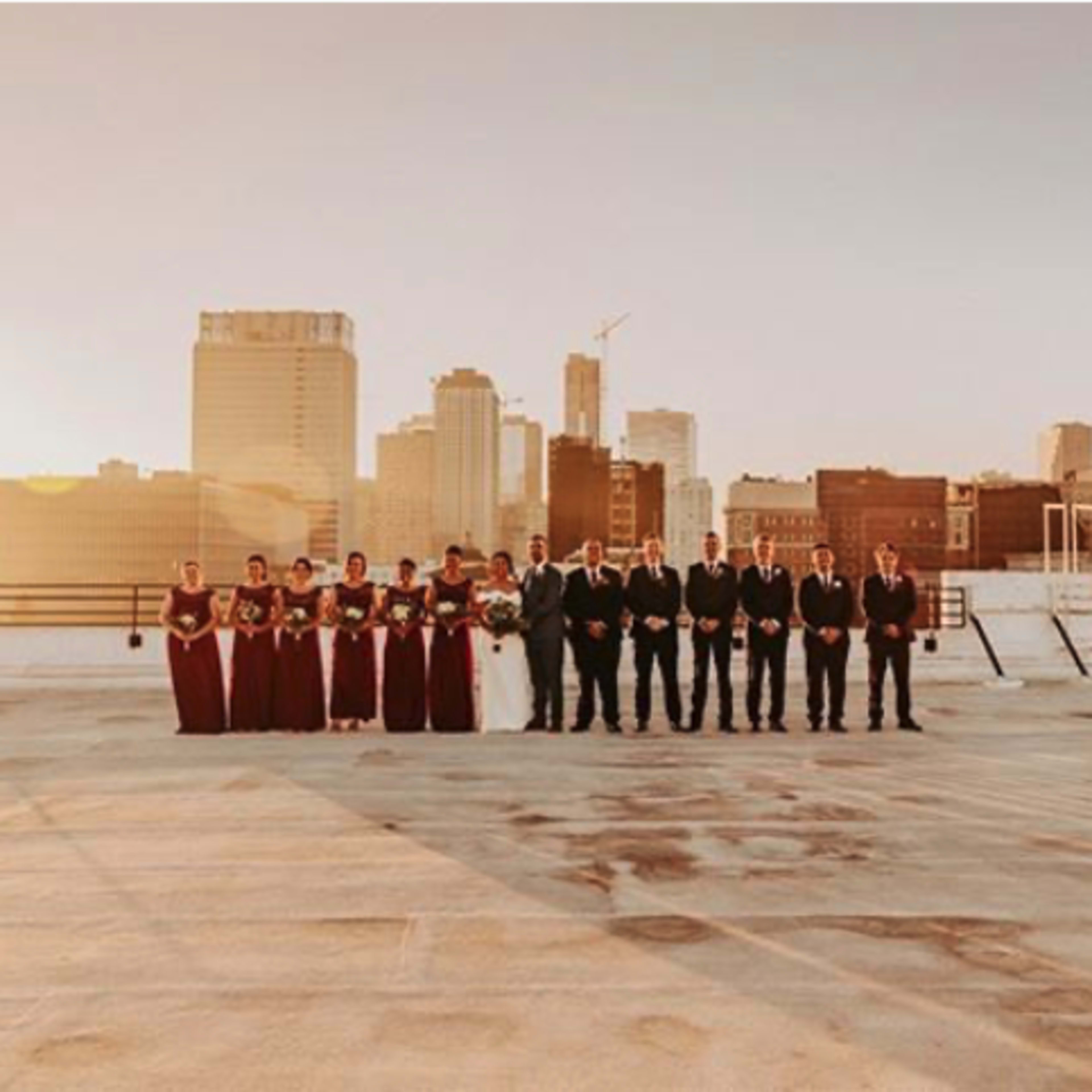 A wedding party in formal attire stands on a rooftop with a city skyline in the background during sunset.