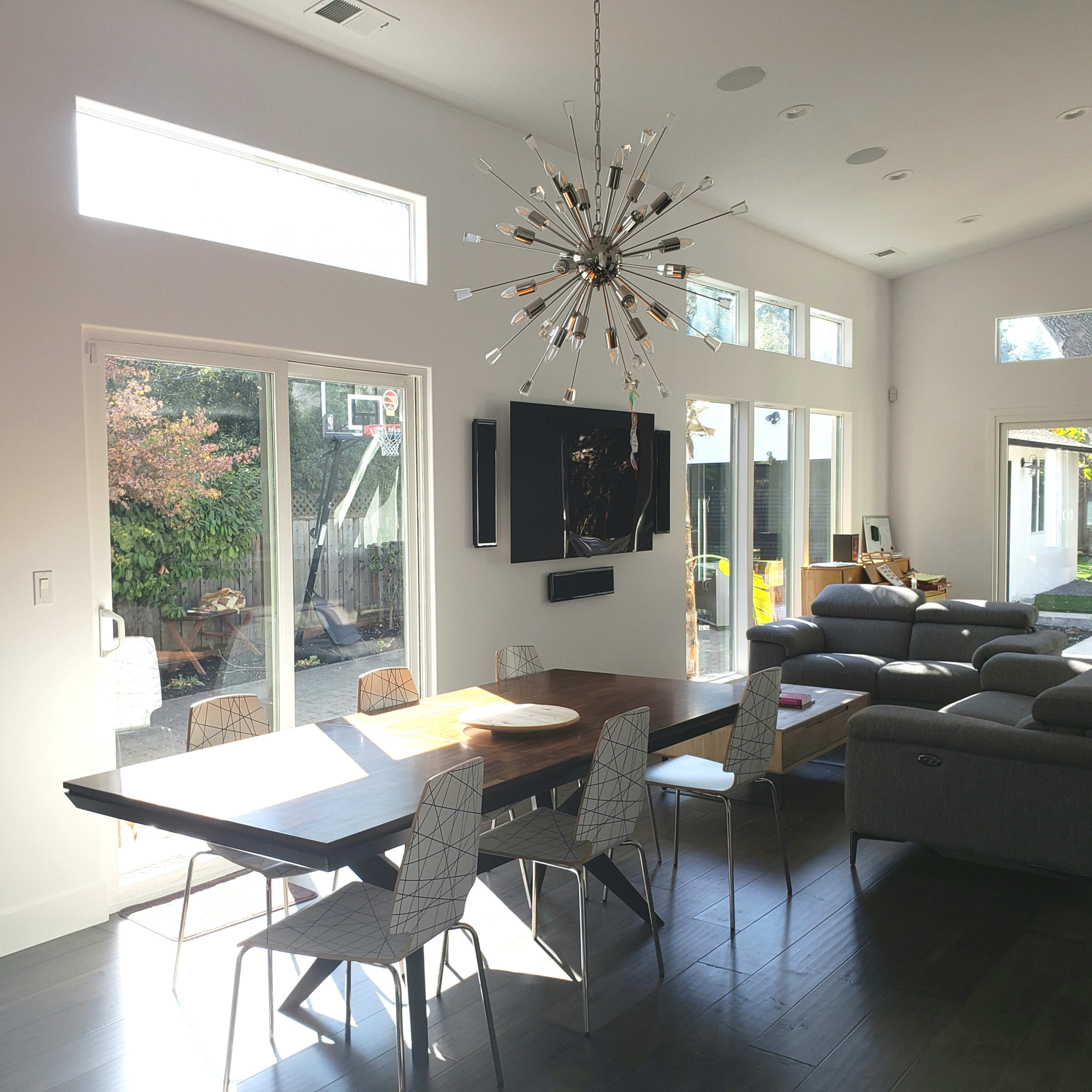 A modern dining area with a large wooden table, sleek chairs, and a starburst chandelier, adjacent to a living space with grey sofas and large windows.