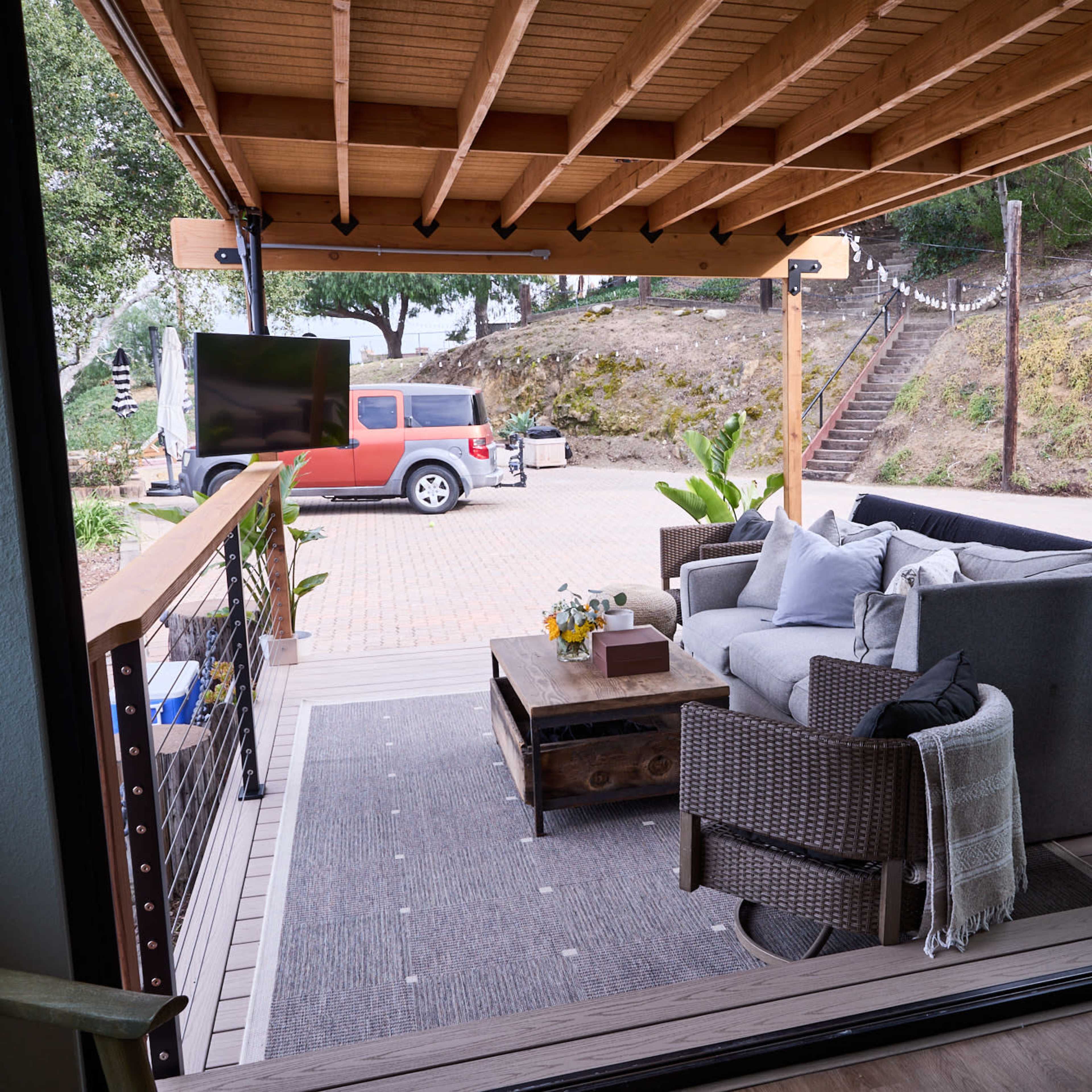 A covered outdoor seating area features a sofa, coffee table, and decorative plants, overlooking a driveway with a parked red vehicle in the background.