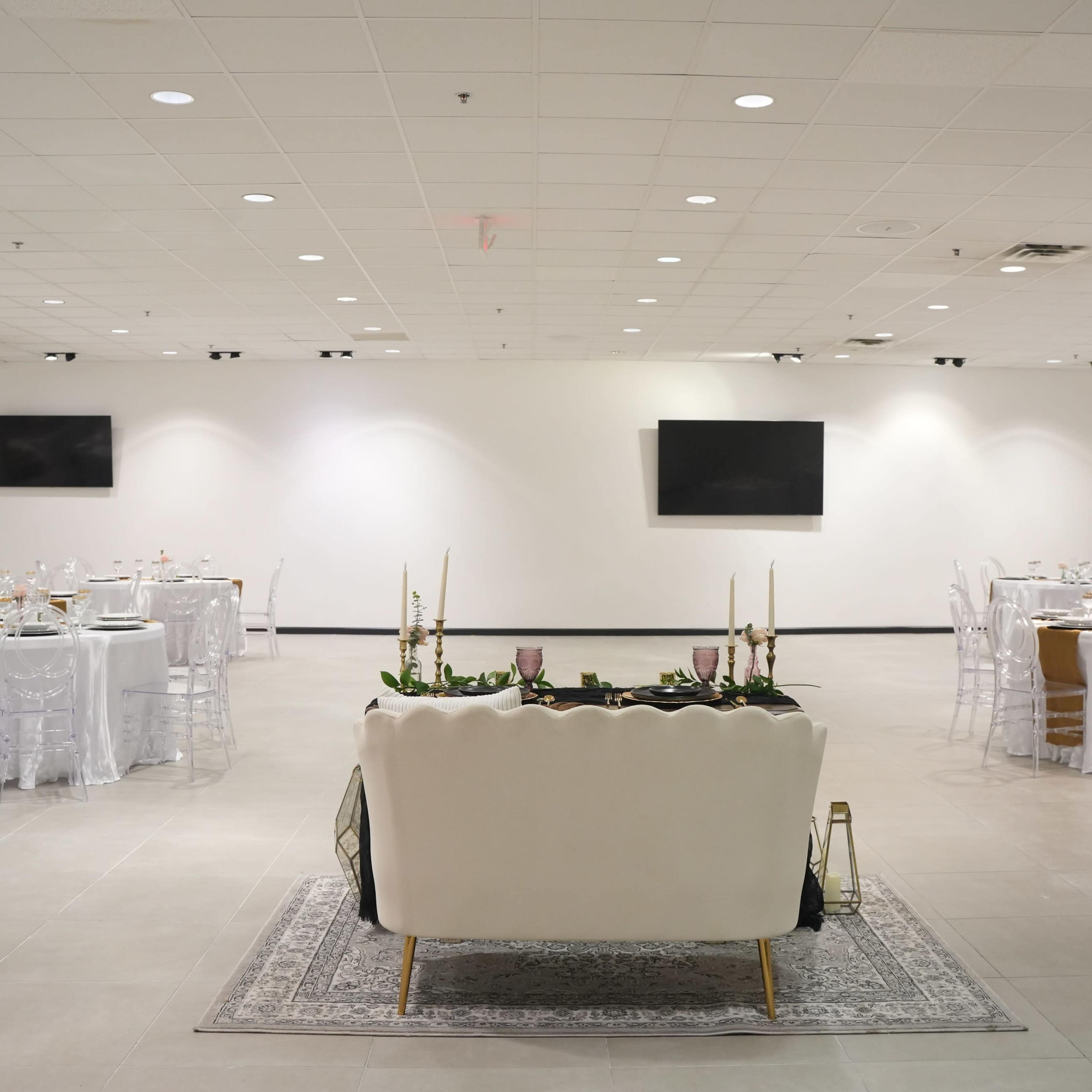 A spacious banquet hall features round tables set for dining and a white couch positioned centrally, with minimal decor and wall-mounted screens.