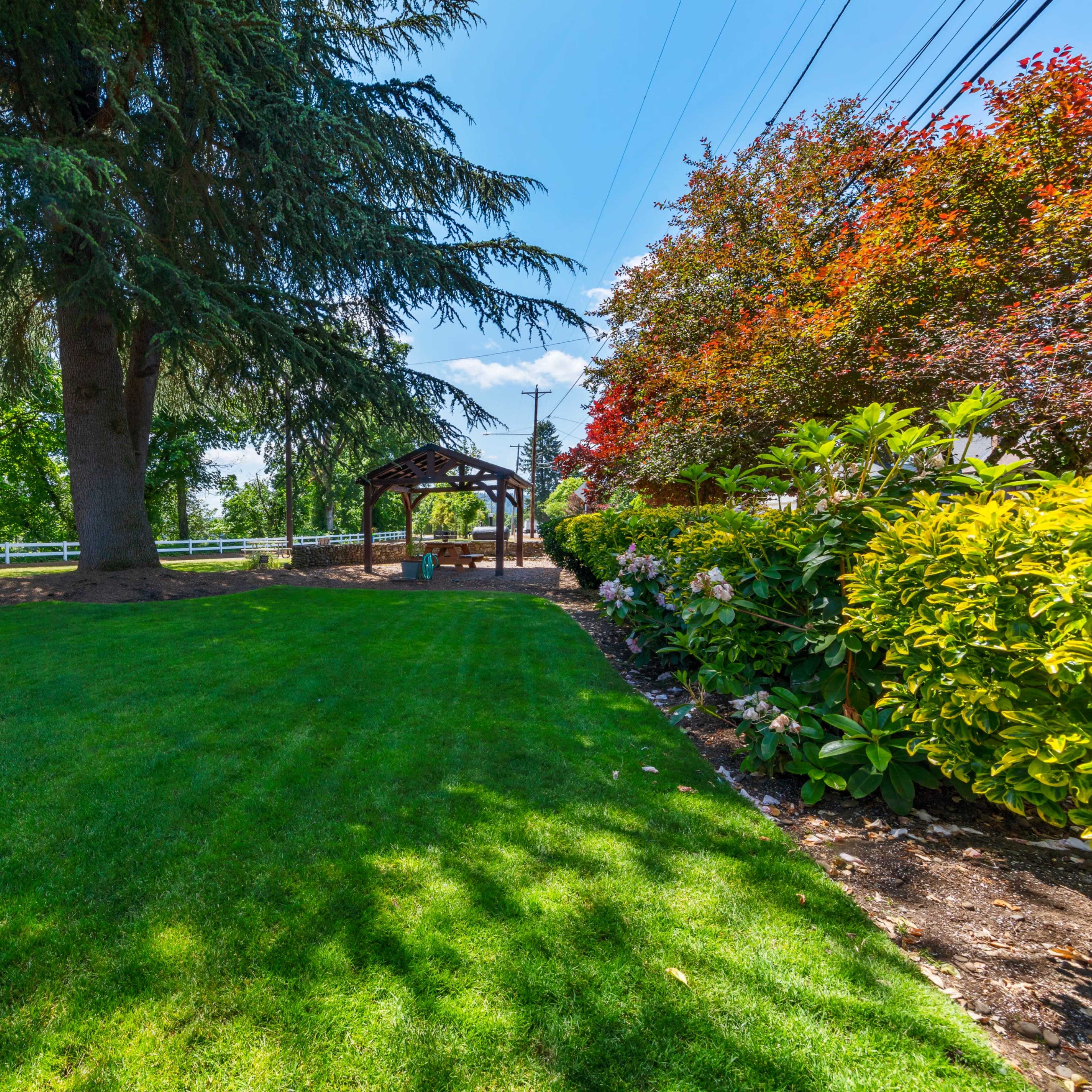 A landscaped garden with neatly trimmed grass, a wooden gazebo, and colorful flowering bushes under a clear blue sky.