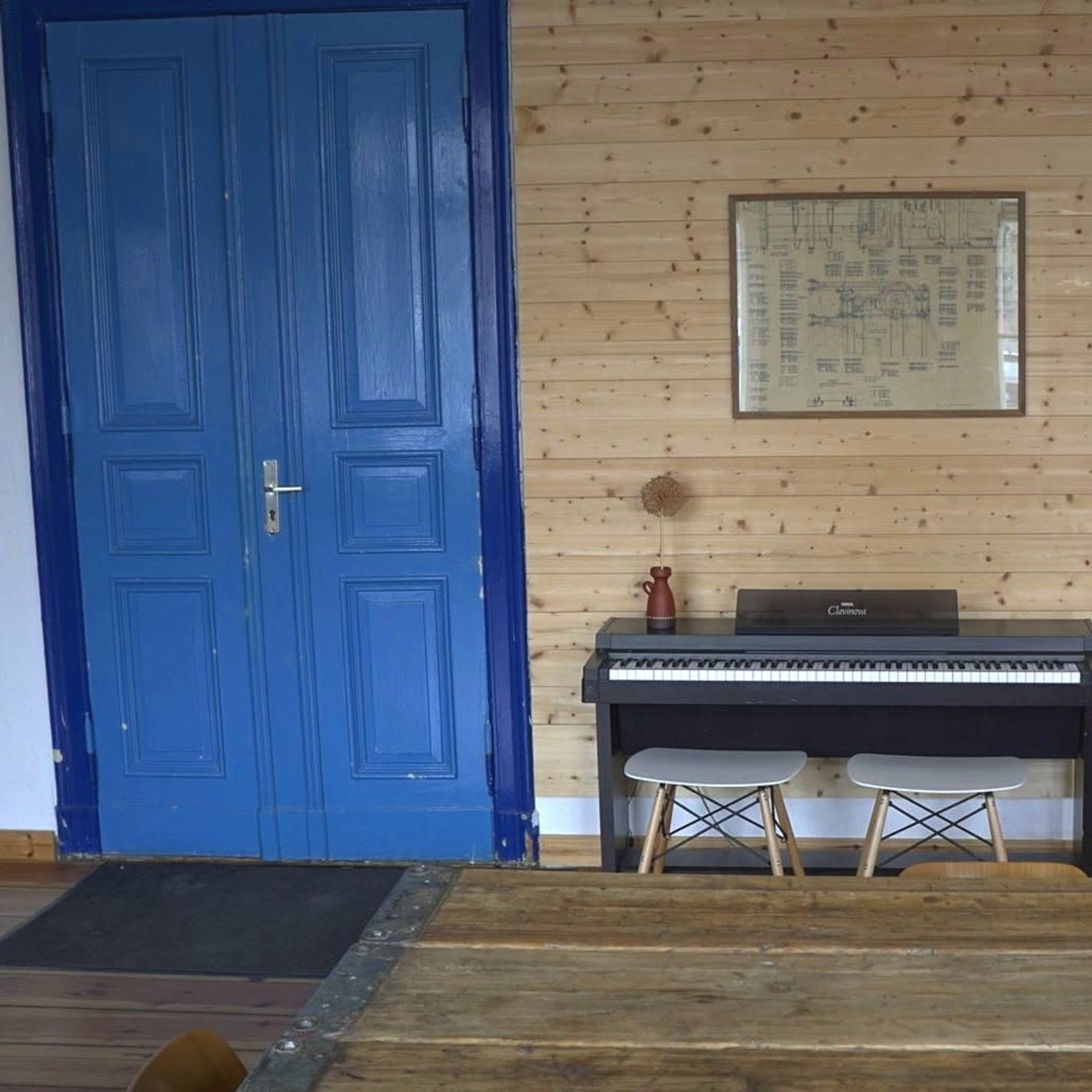 A black piano sits against a wooden wall with a blue door nearby and a framed piece of art above it.