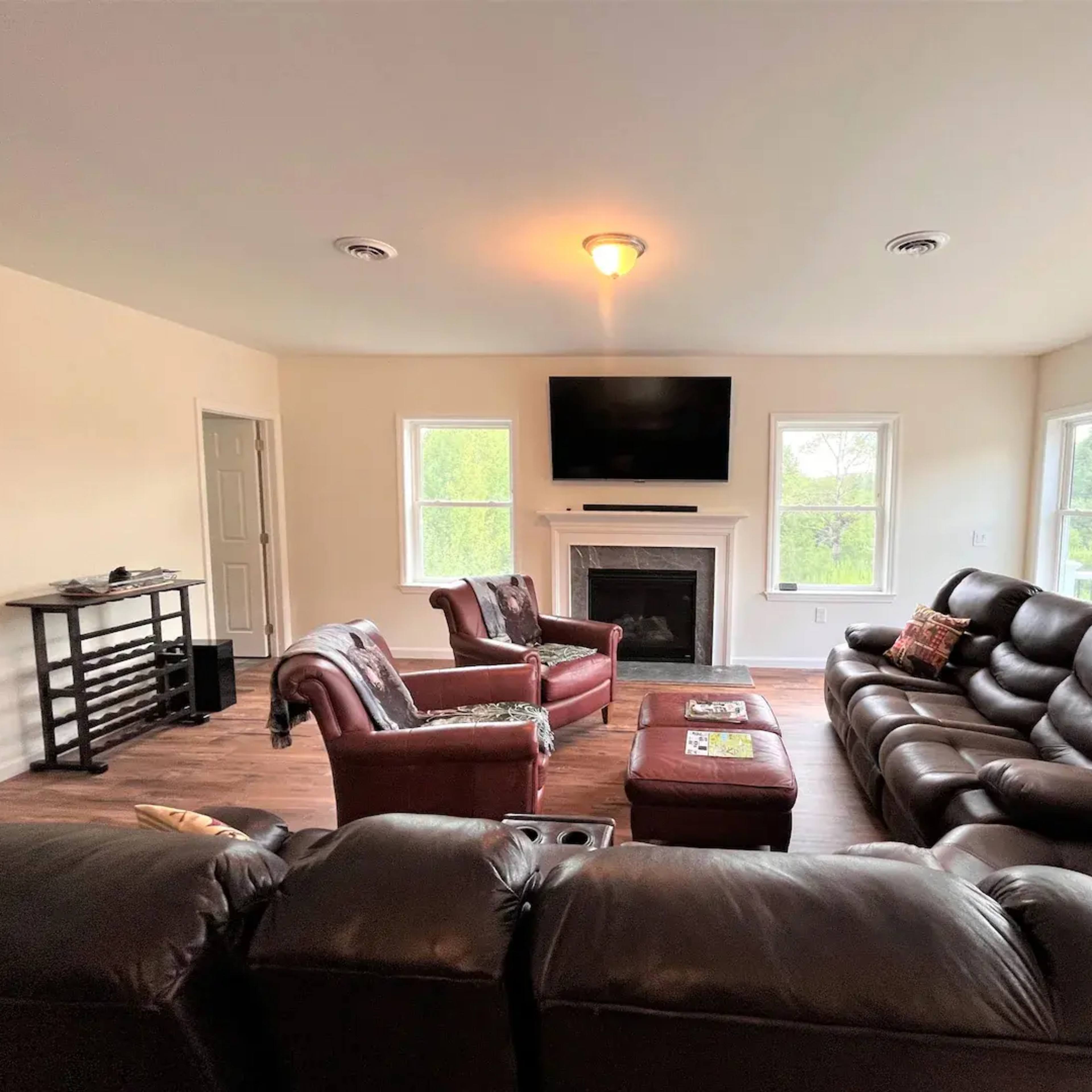 The living room features a dark leather sectional sofa, two matching armchairs, a coffee table, a media console, and a wall-mounted television, with large windows providing views of the outdoors.