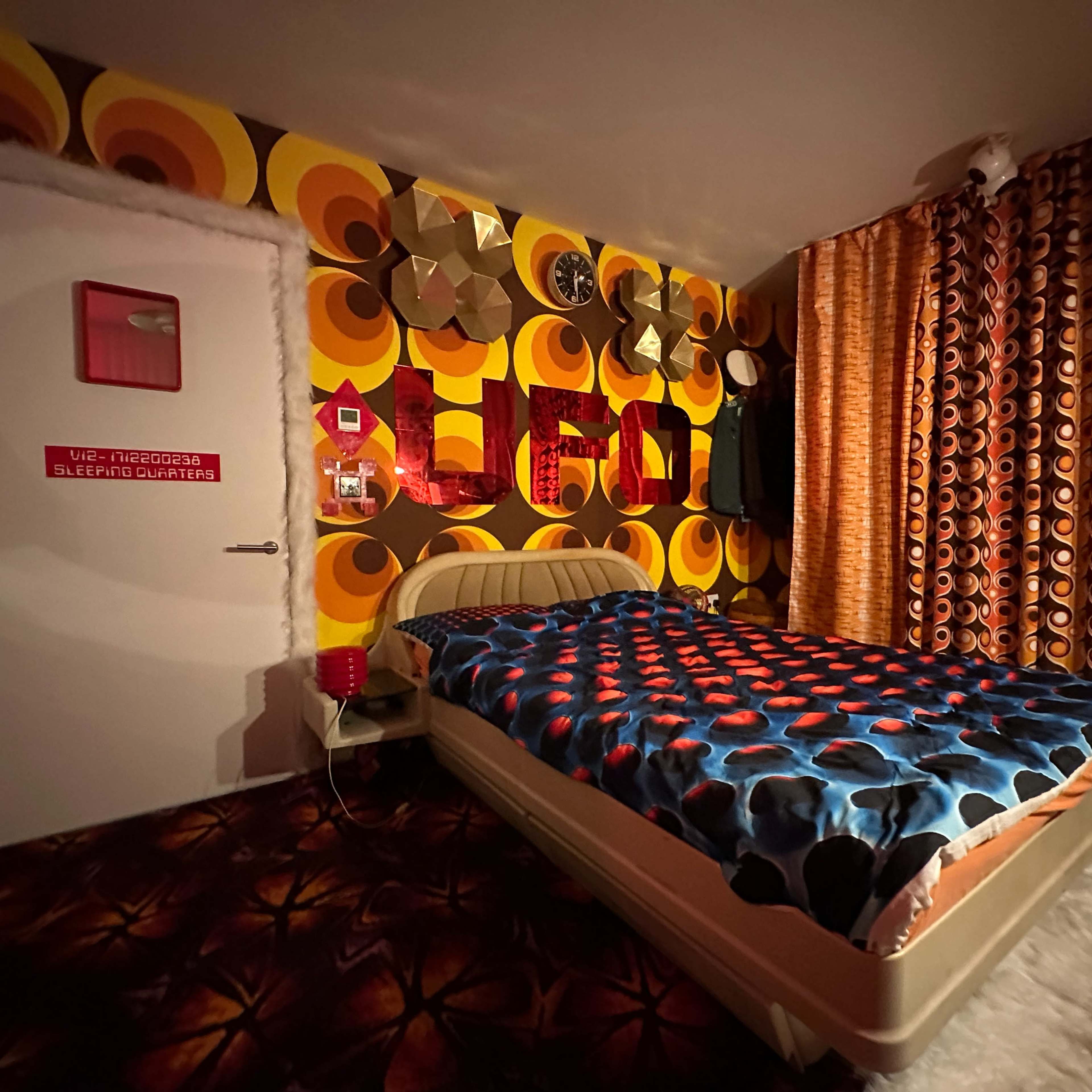 A retro-themed bedroom featuring bold patterned wallpaper, a patterned bedspread, and vibrant curtains.