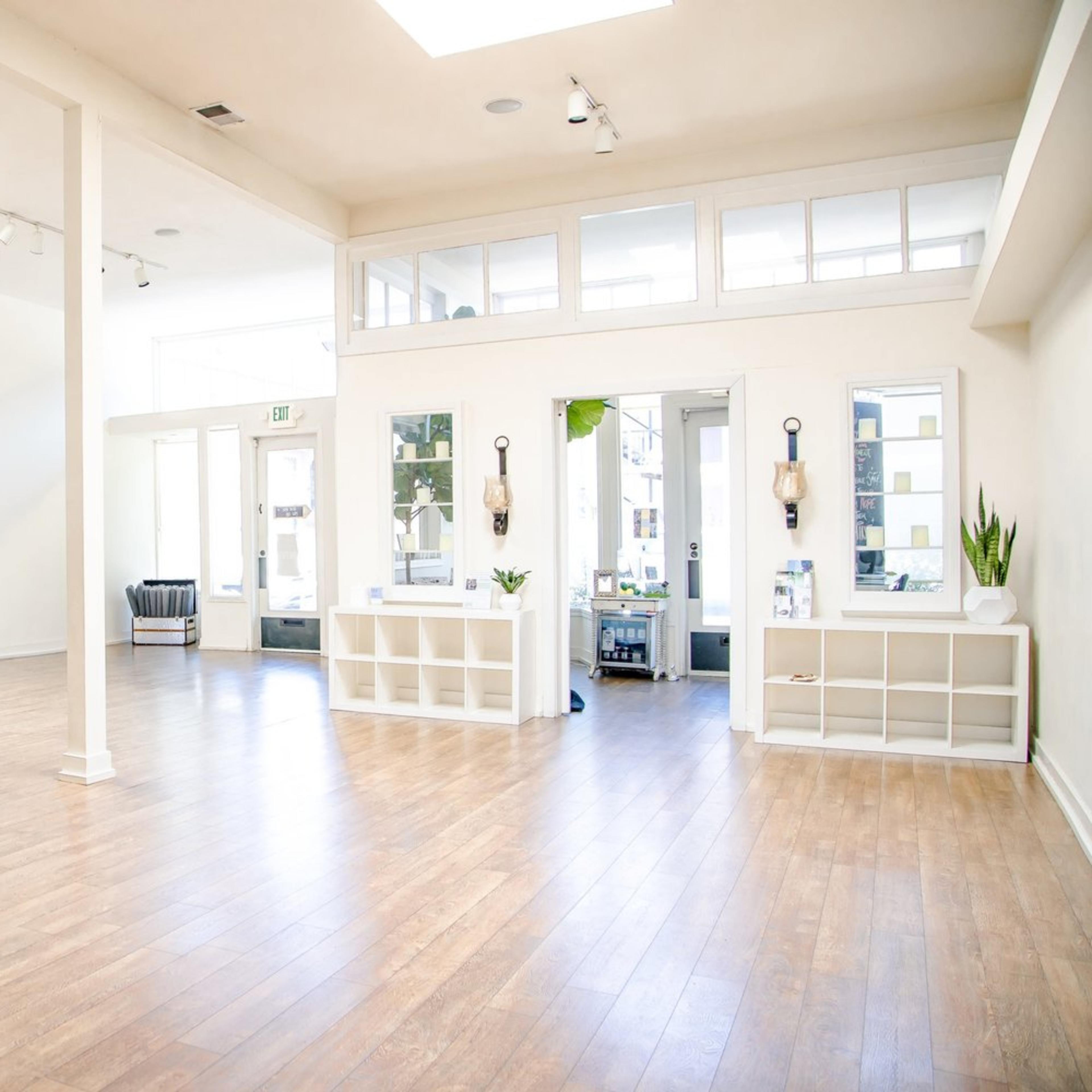 A spacious, empty room with wooden flooring, featuring large windows, minimal furnishings, and plants on display.