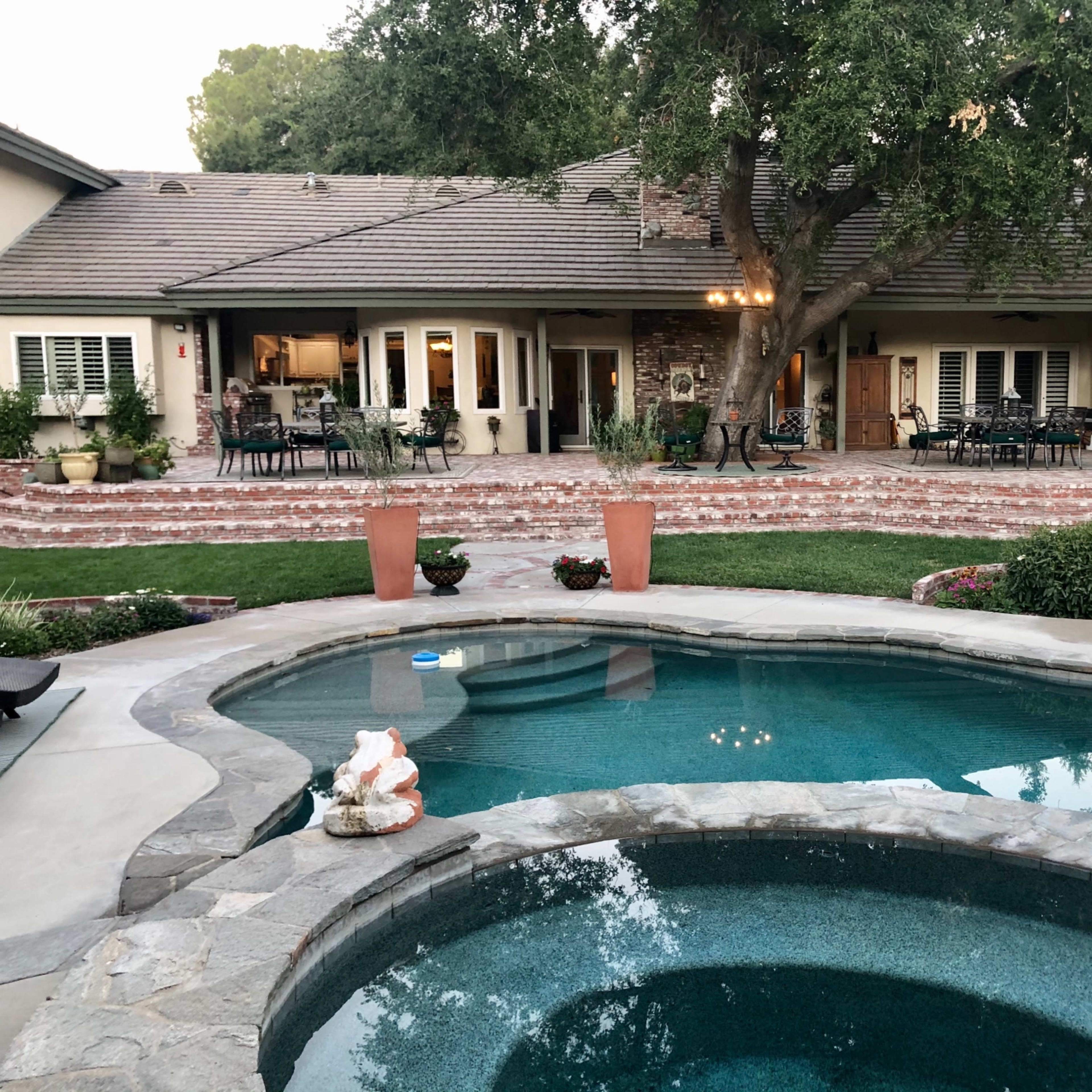 A landscaped backyard with a swimming pool, a spa, and a large house in the background surrounded by greenery and patio furniture.
