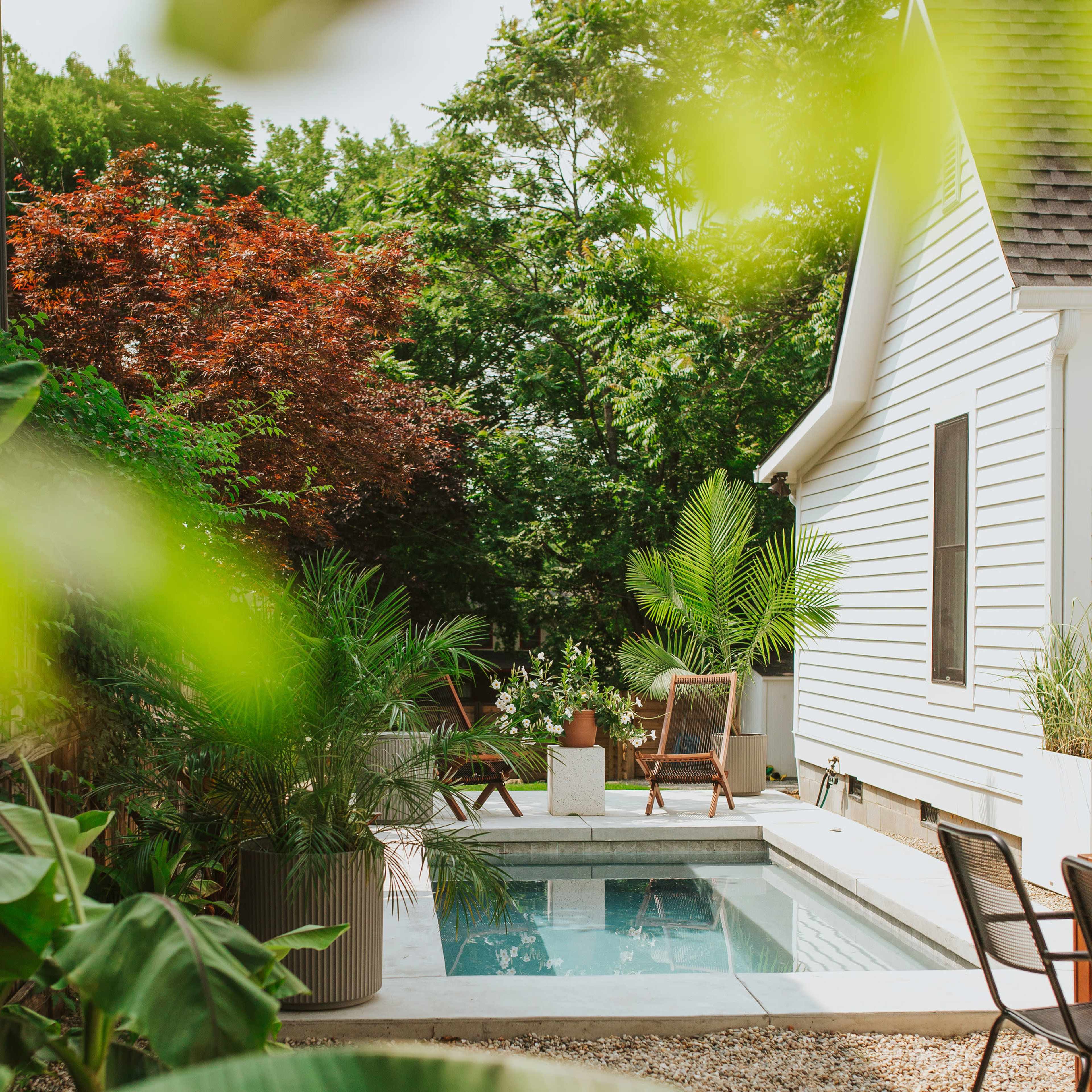 A small, rectangular pool sits beside a white house, surrounded by greenery and outdoor furniture in a landscaped yard.