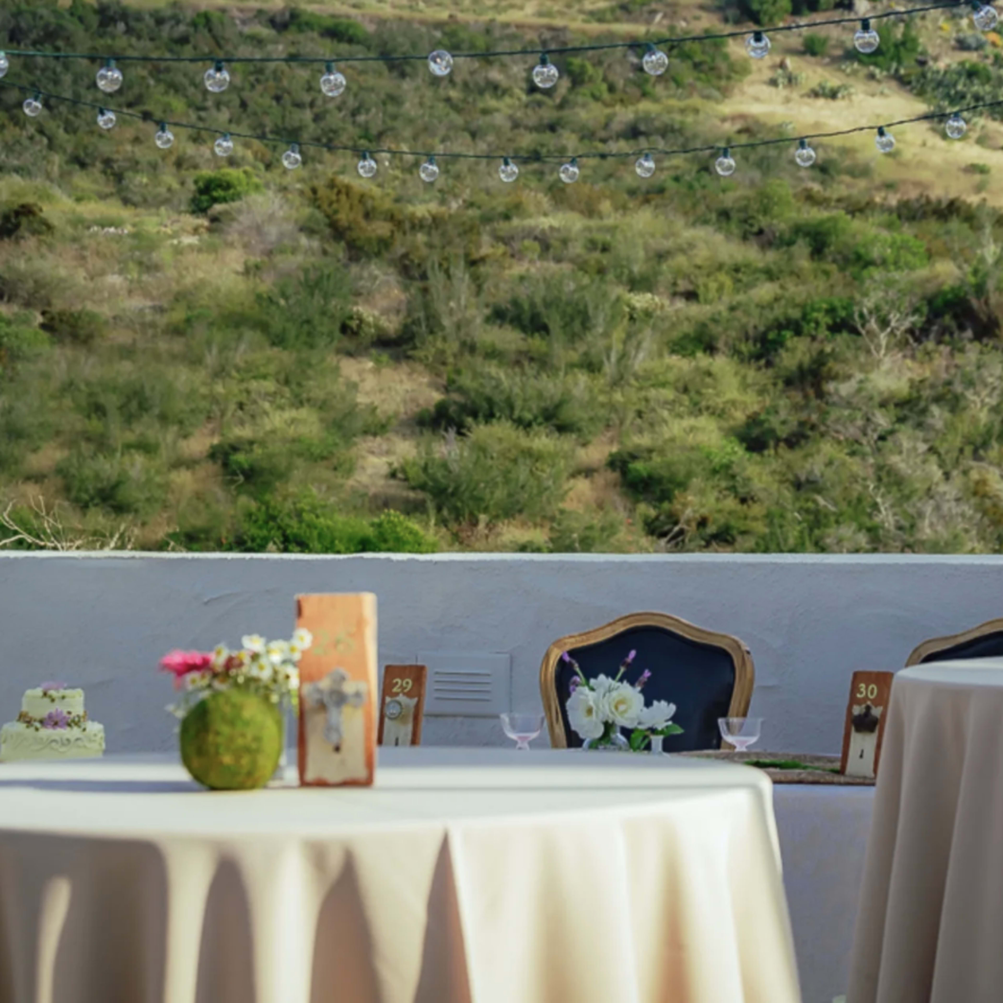 A dinner setting with round tables covered in beige tablecloths, featuring floral centerpieces and decorative number signs, overlooks a lush green hillside.