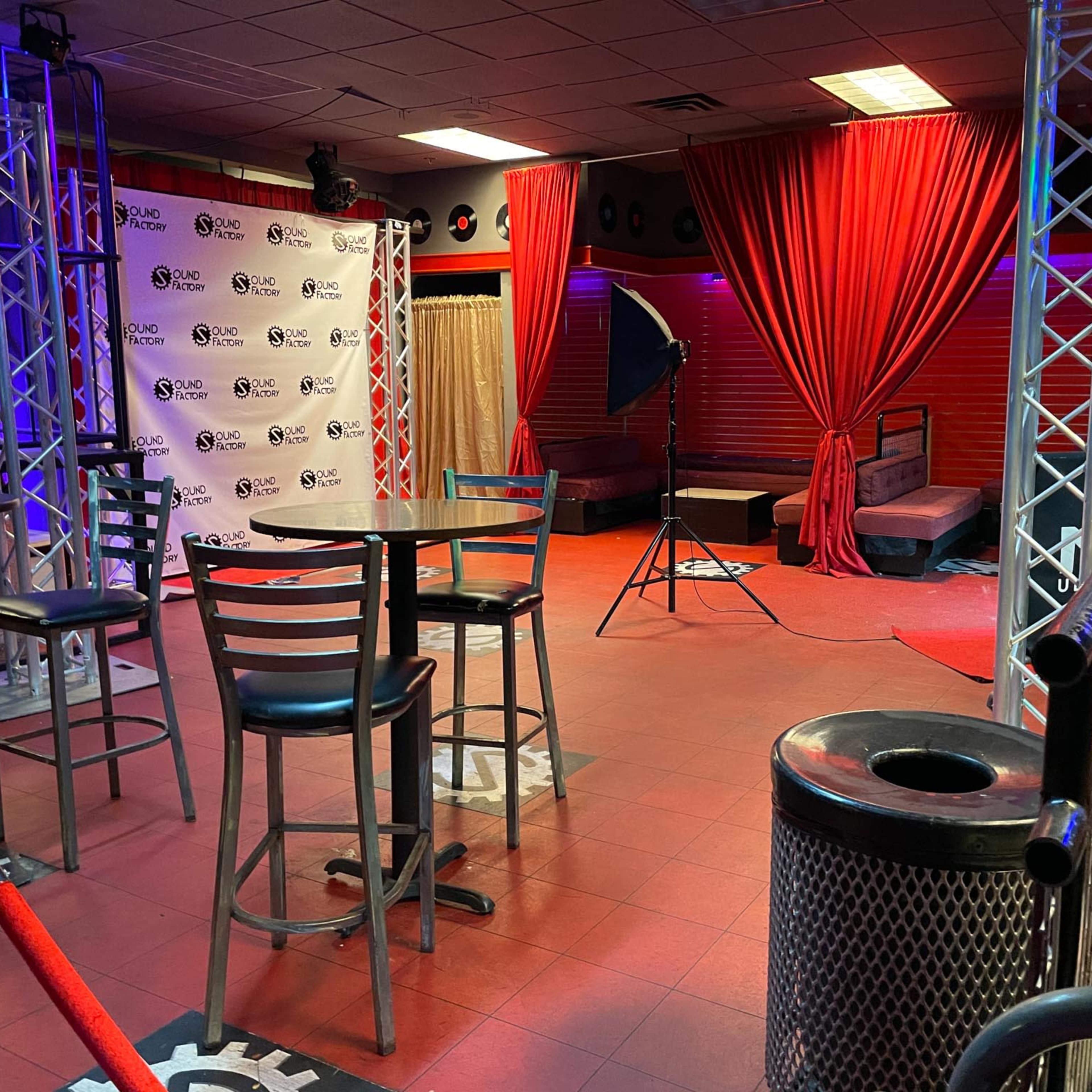 A dimly lit bar area with red curtains, empty tables, and a photography setup in the corner.