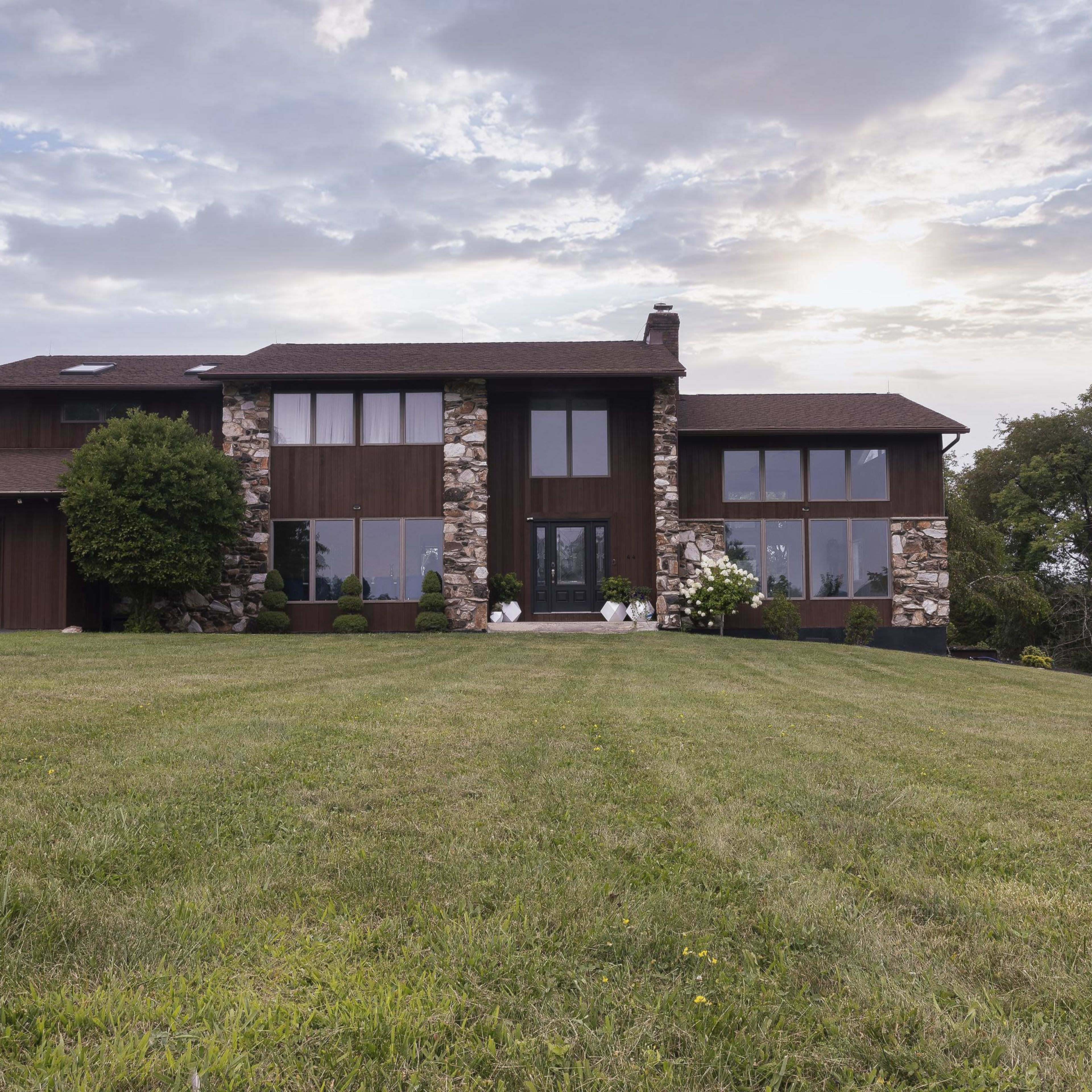 A modern house with a stone facade and large windows sits on a green lawn under a cloudy sky.