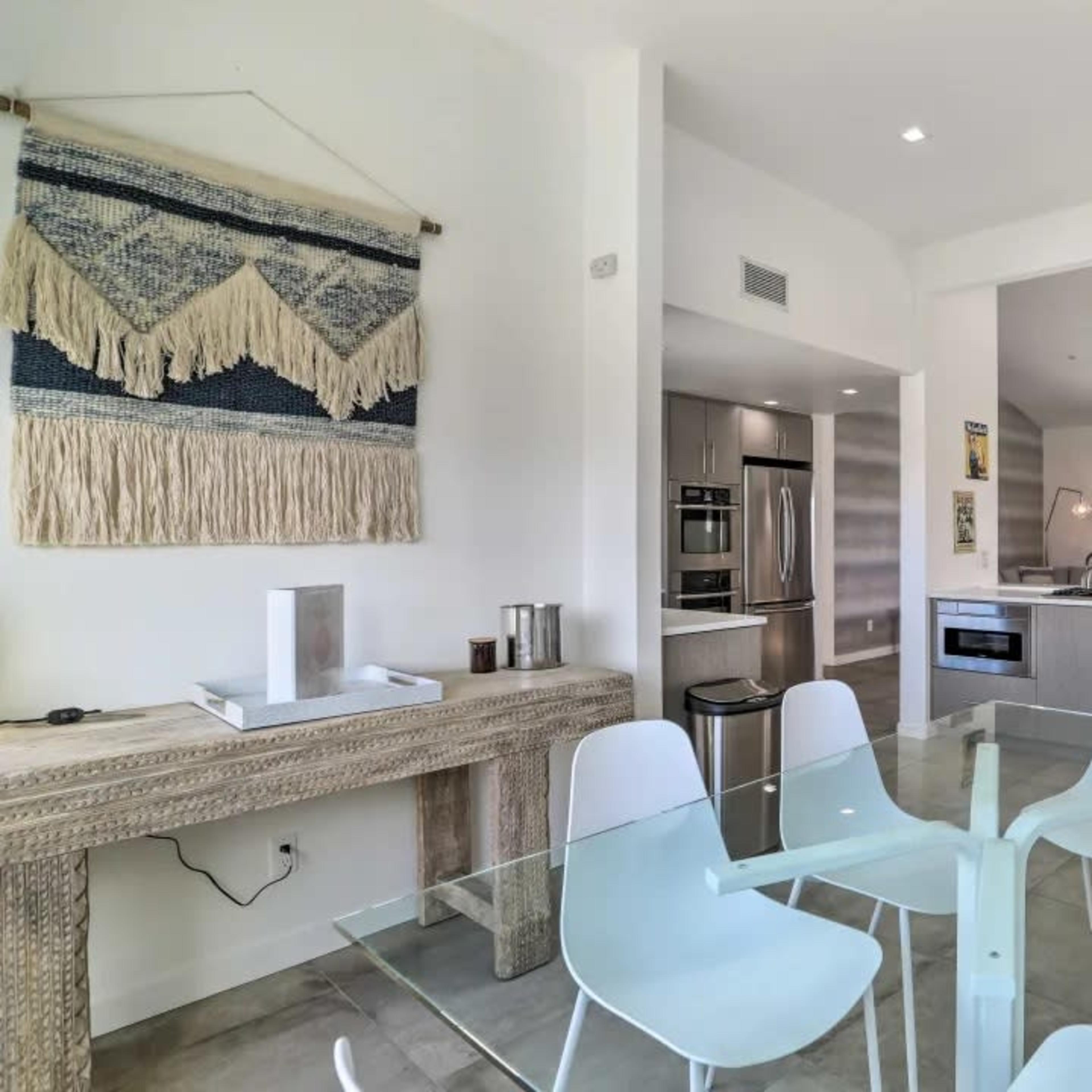 A modern kitchen and dining area features a glass table surrounded by white chairs, with a wooden console table and a stylish wall hanging.