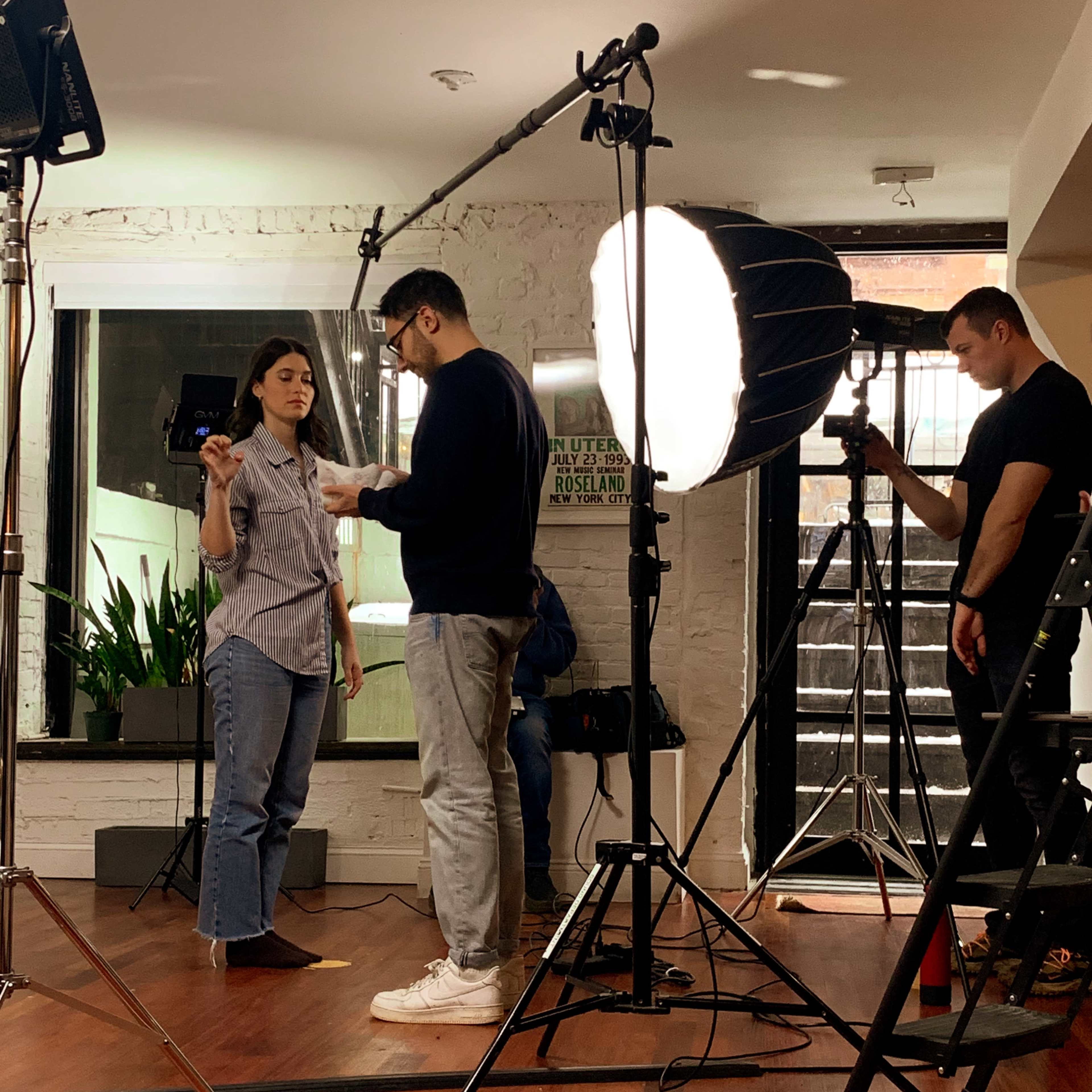 A woman stands in a well-lit room with a camera setup, while a man adjusts equipment nearby and another person operates a camera.