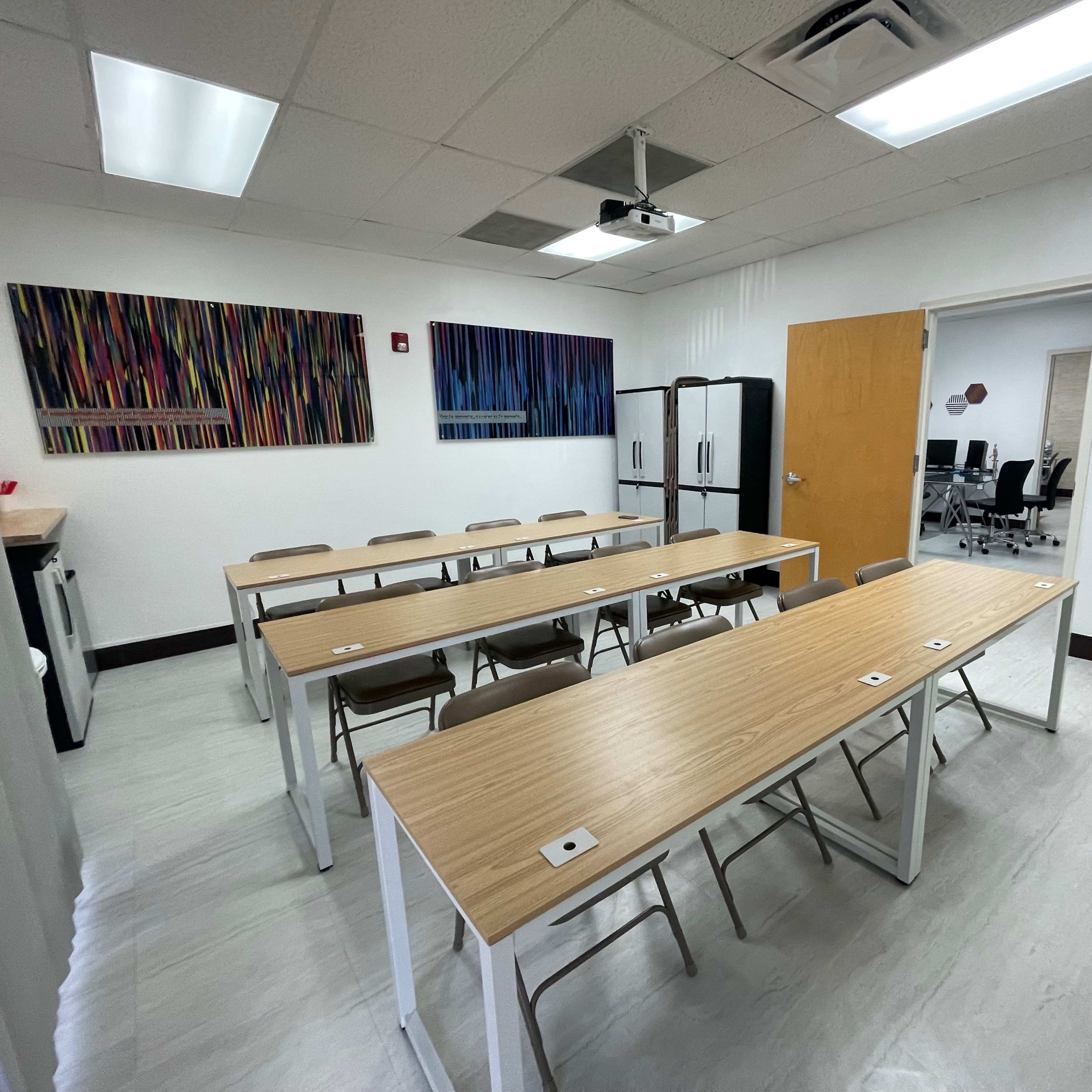 A bright classroom with wooden tables arranged in rows, folding chairs, a refrigerator, and colorful wall art.
