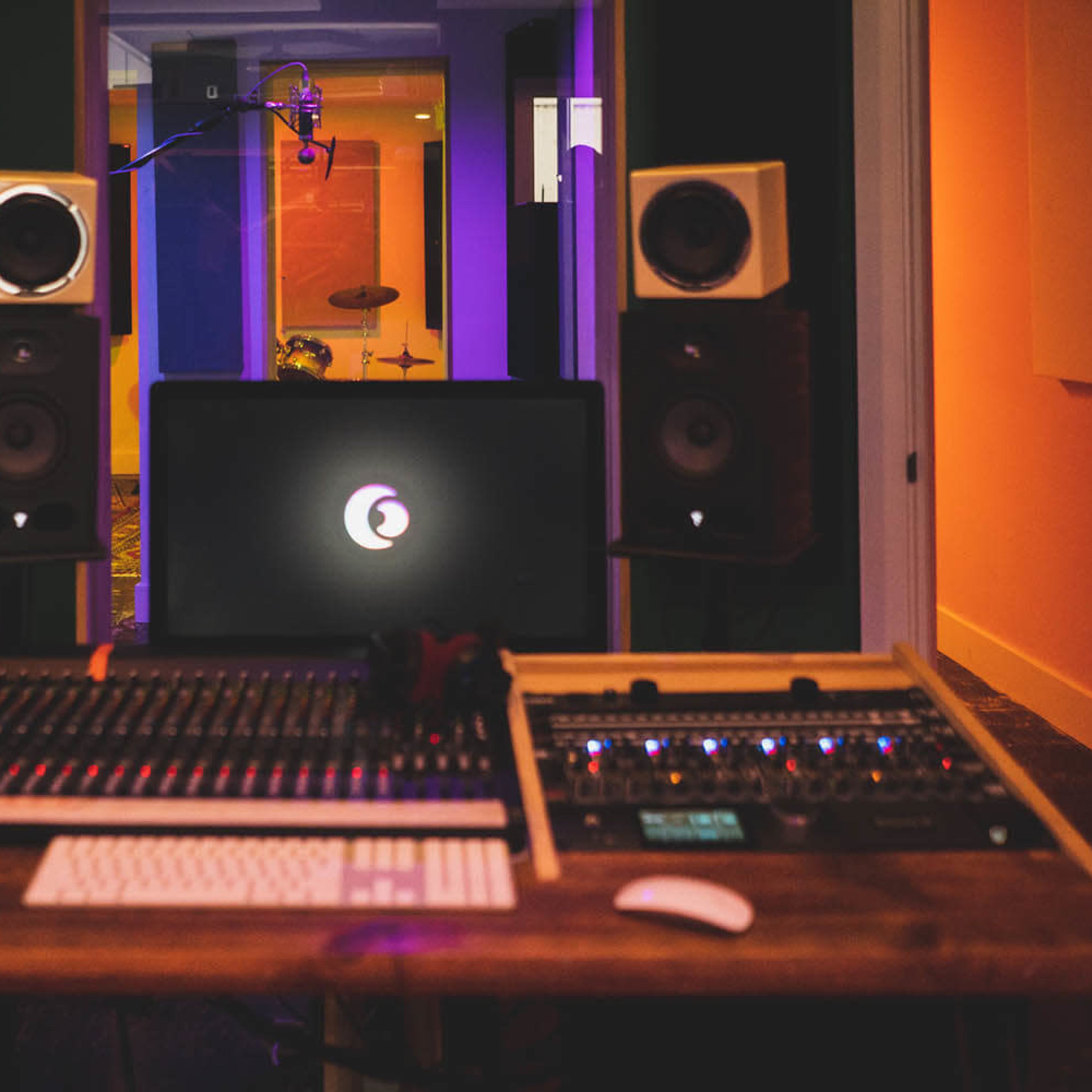 A recording studio setup features a large mixing console, computer, and studio monitors, with a microphone and drum set visible in the background.