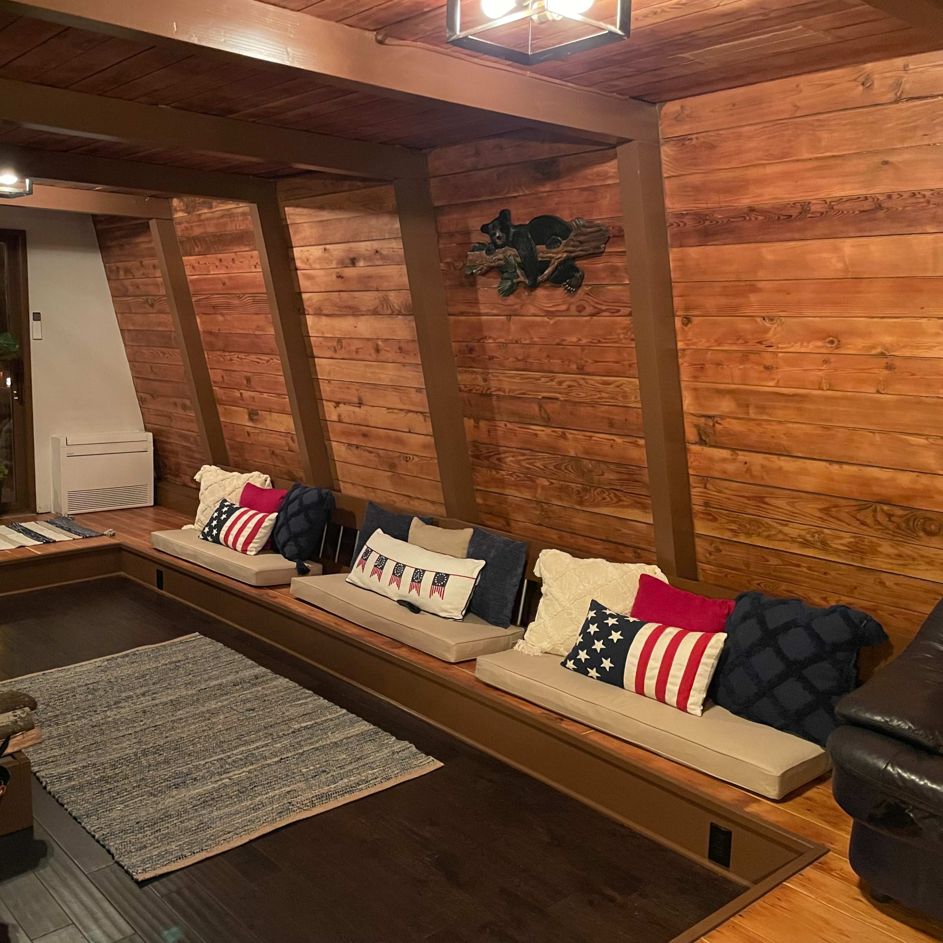 A cozy interior with wooden walls, a long seating area filled with decorative pillows, and a staircase in the background.