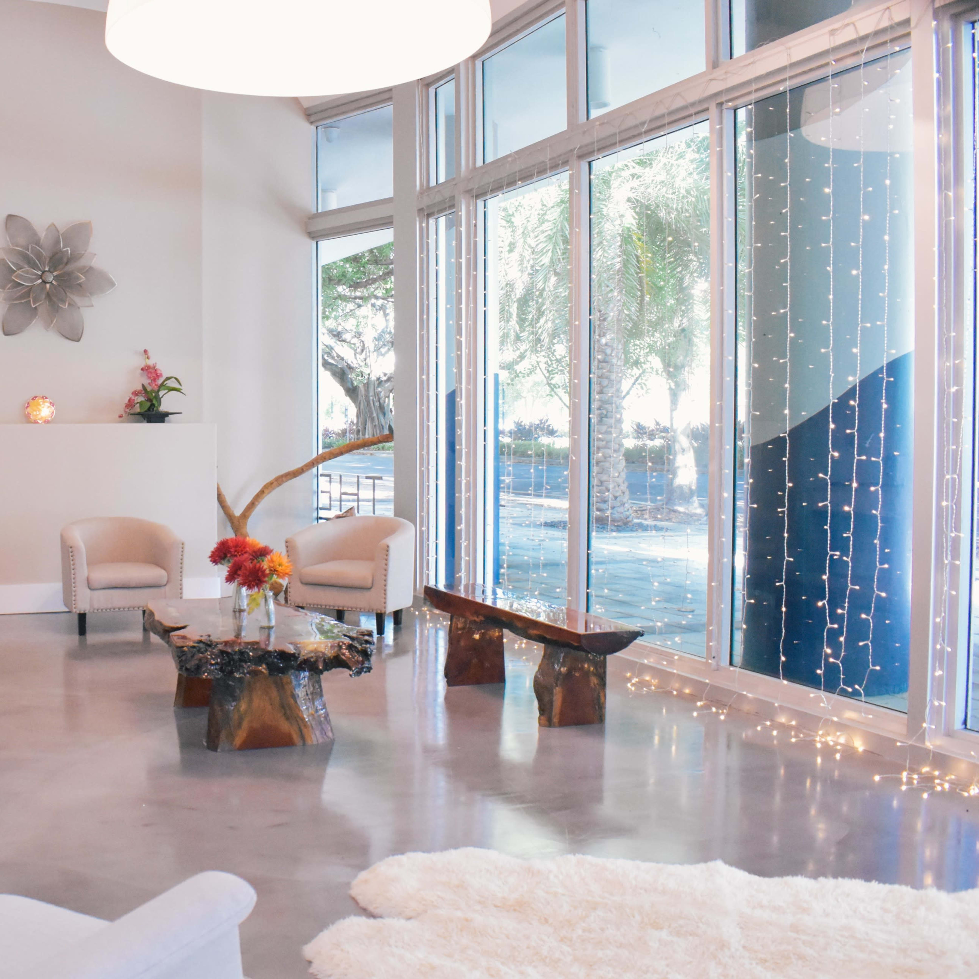 The scene features a modern living space with large windows, a light-colored sofa and chairs, a wooden coffee table, and decorative plants.